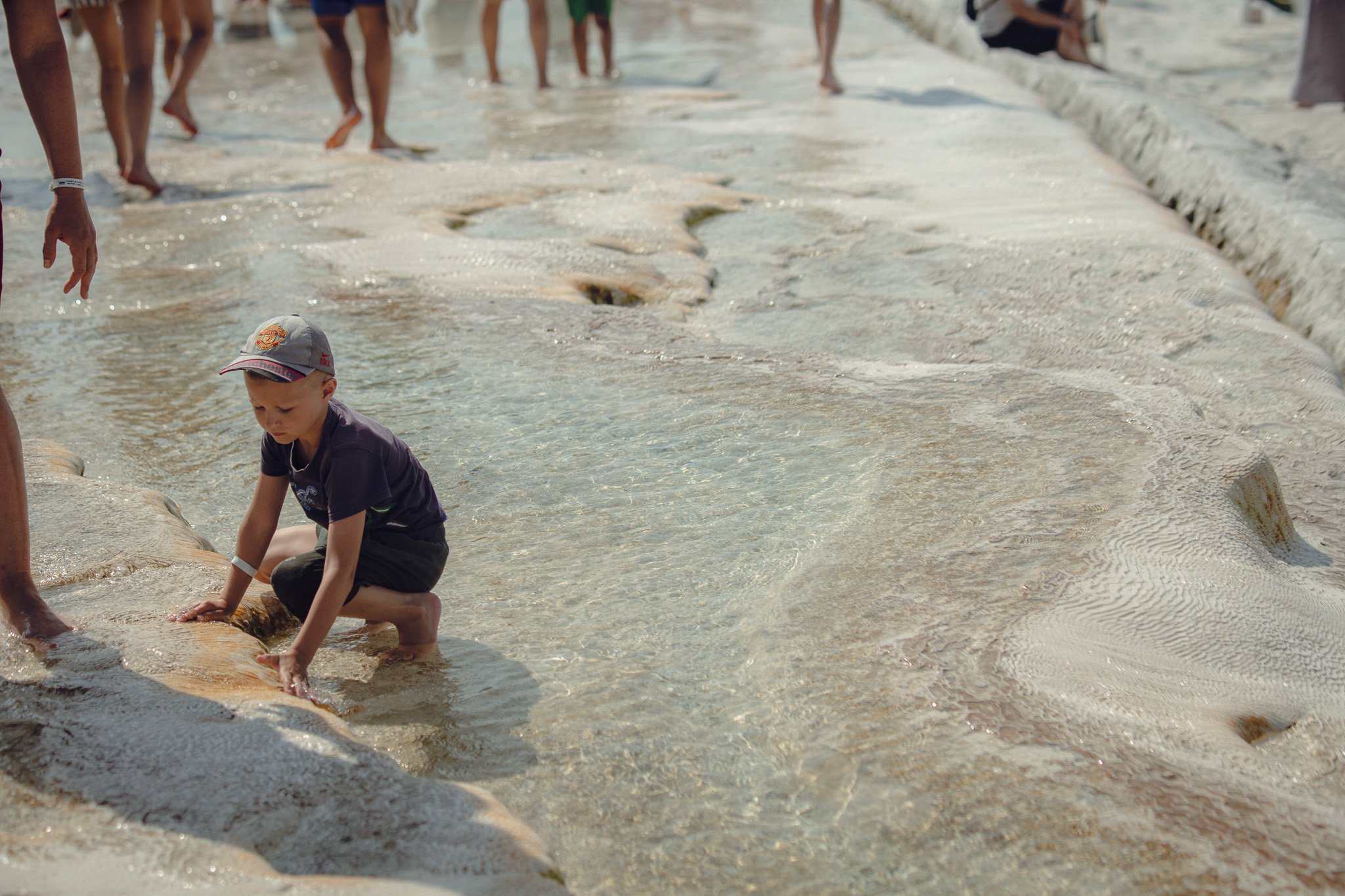 A young boy crouches in shallow, clear water at the beach, wearing a baseball cap and a dark t-shirt, surrounded by adults and other children in the background.