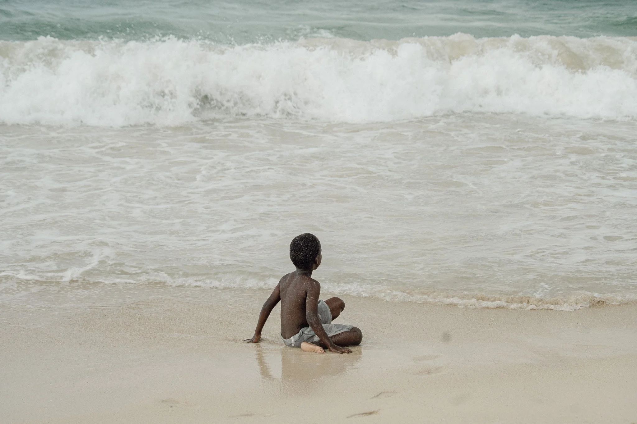 A young boy sitting on the sandy beach looking at the ocean waves.