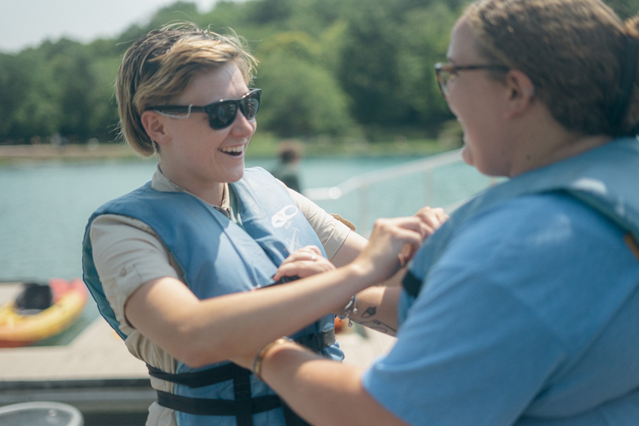 Two women smiling and holding hands on a boat near a lake with green trees in the background.