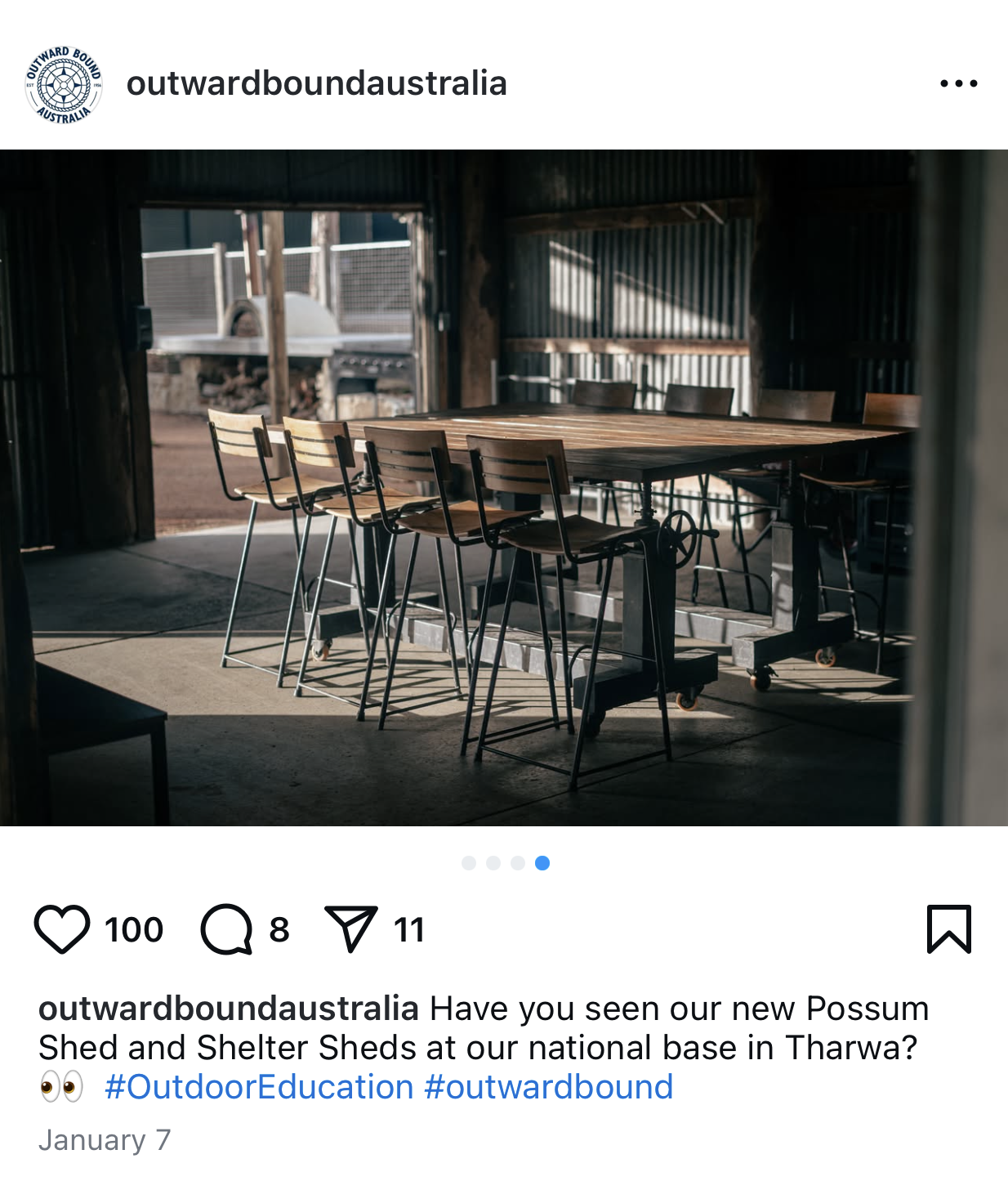 A rustic indoor setting with a large wooden table surrounded by high chairs, inside a shed with corrugated metal walls and a partially open door showing an outdoor area with a grill and firewood.