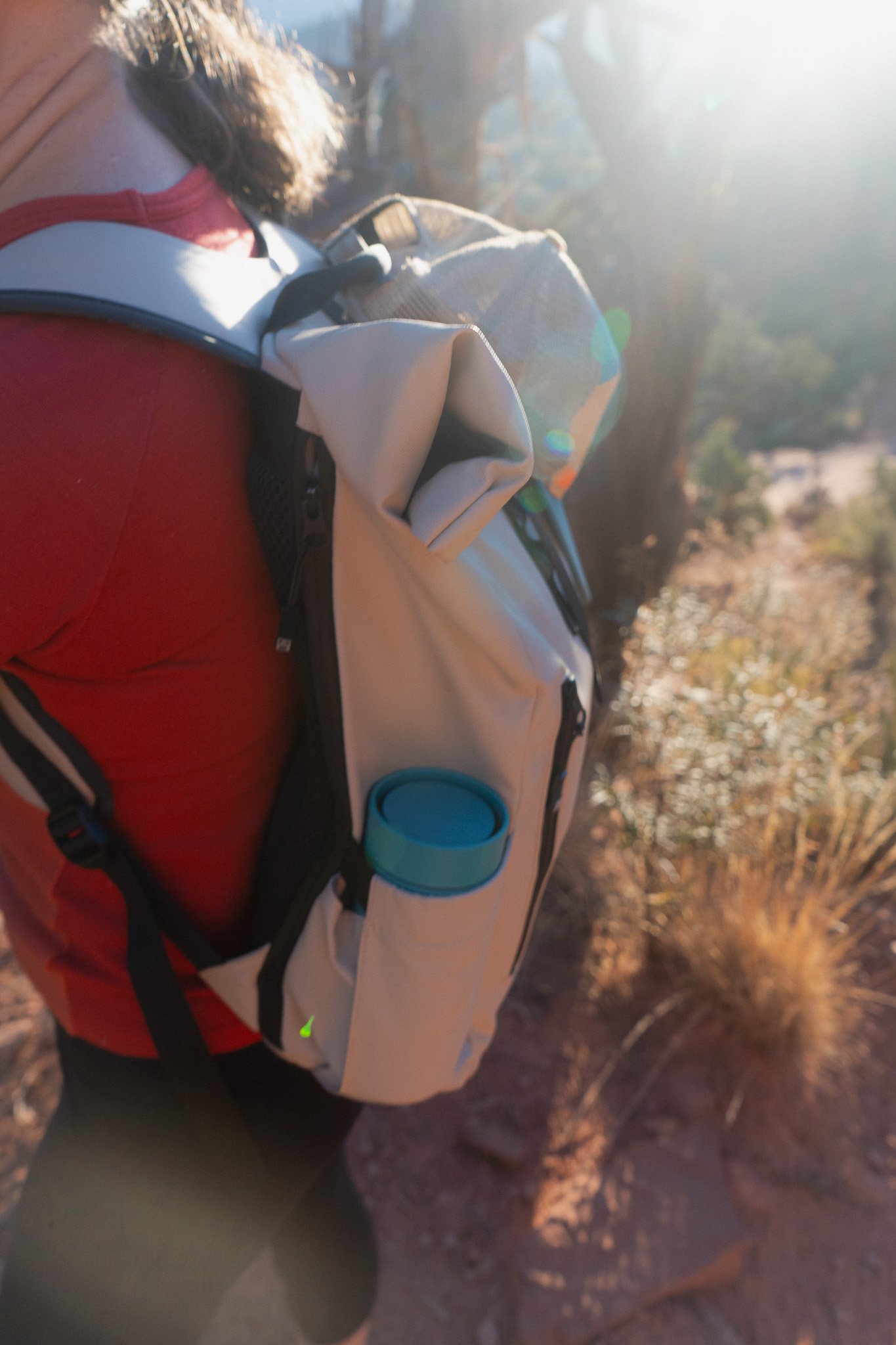 A person wearing a red shirt hiking on a trail during sunset, carrying a beige backpack with a blue collapsible cup attached.