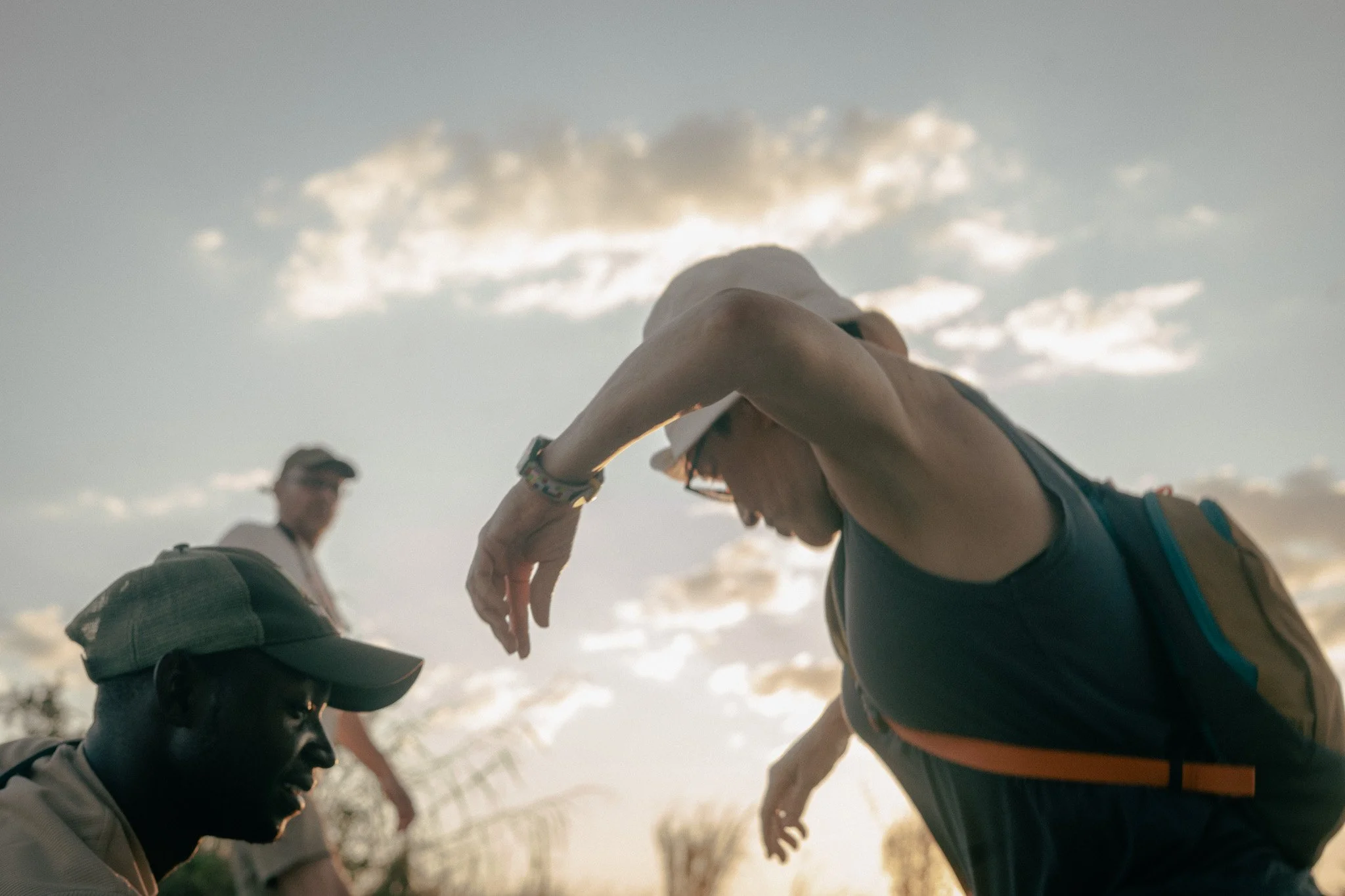 A young person with a backpack bending forward, with a woman nearby and a man in the background, against a cloudy sky during sunset.