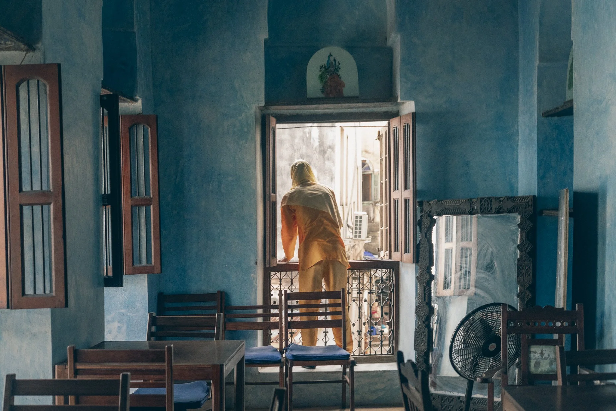Person in beige clothing standing at open window in a room with blue walls and wooden furniture