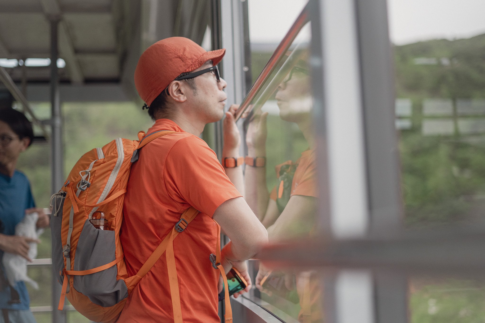 A man wearing an orange t-shirt, orange backpack, orange cap, and glasses looks out the window of a bus or train, with his hand resting on the glass and his reflection visible.