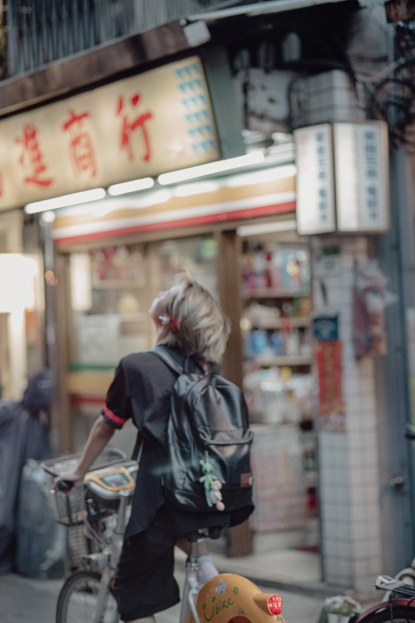 A person with short hair riding a yellow and white bicycle past a shop with Asian signage in an urban setting.