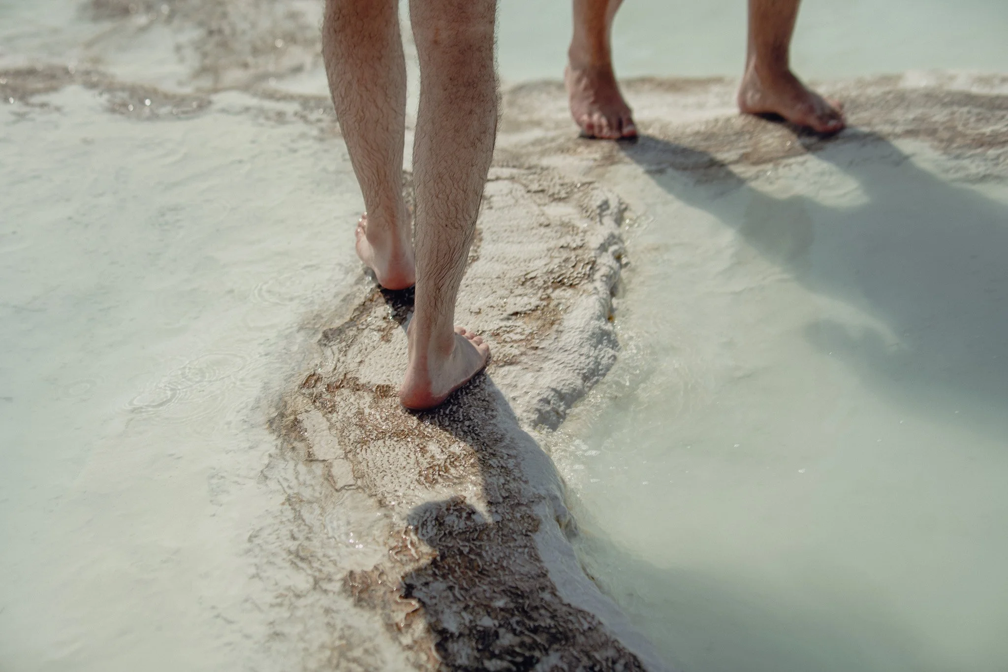 Close-up of two people walking barefoot on wet sand near the ocean during daytime.