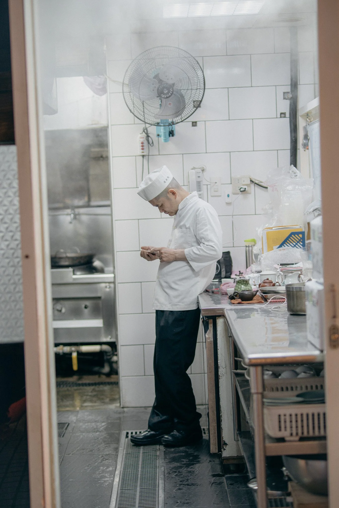 A chef in a white uniform and hat is standing in a kitchen, looking at his phone.