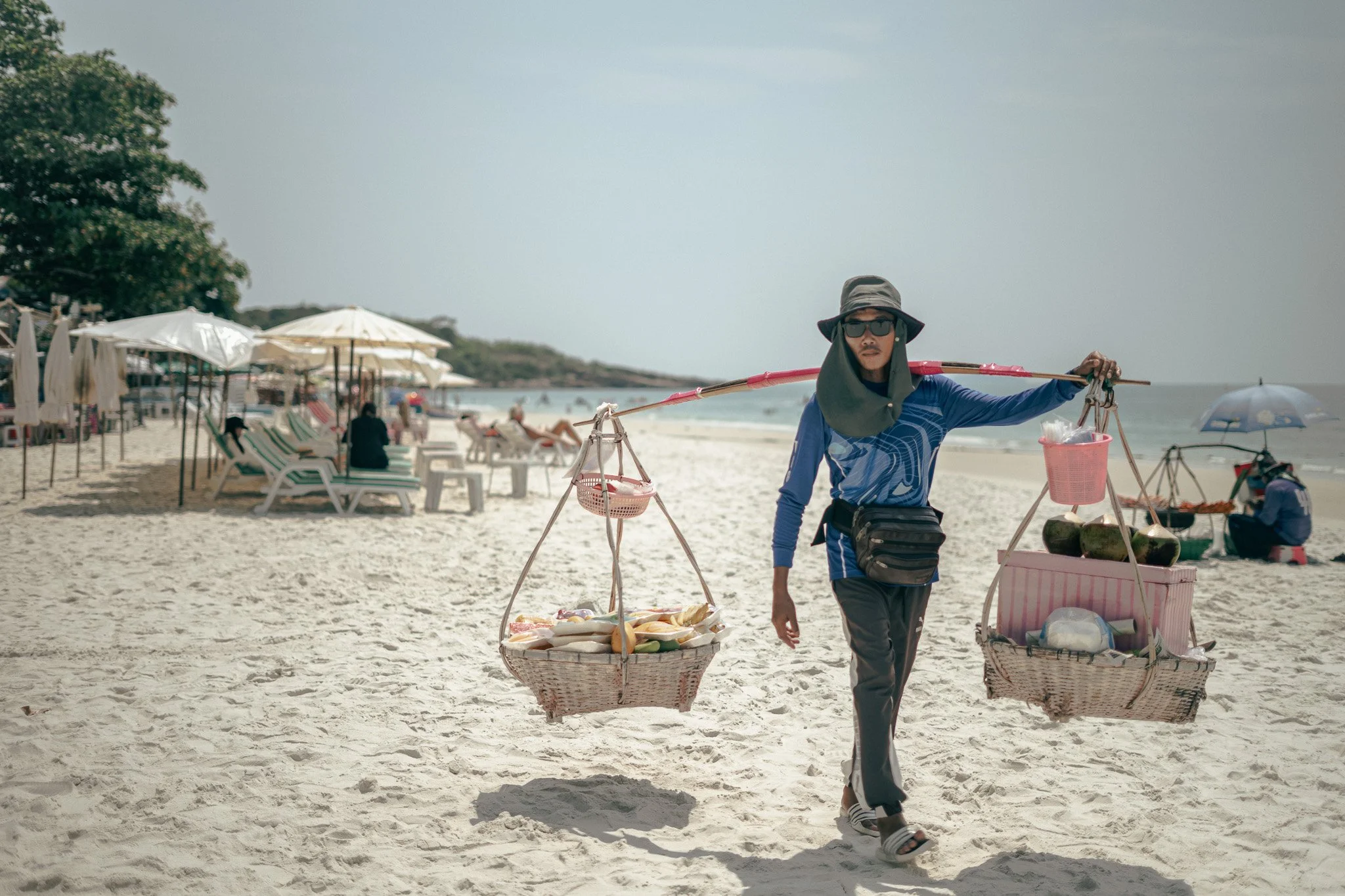 A street vendor walking on the beach carrying baskets with various goods balanced on a pole across his shoulders, with beach umbrellas and loungers in the background.