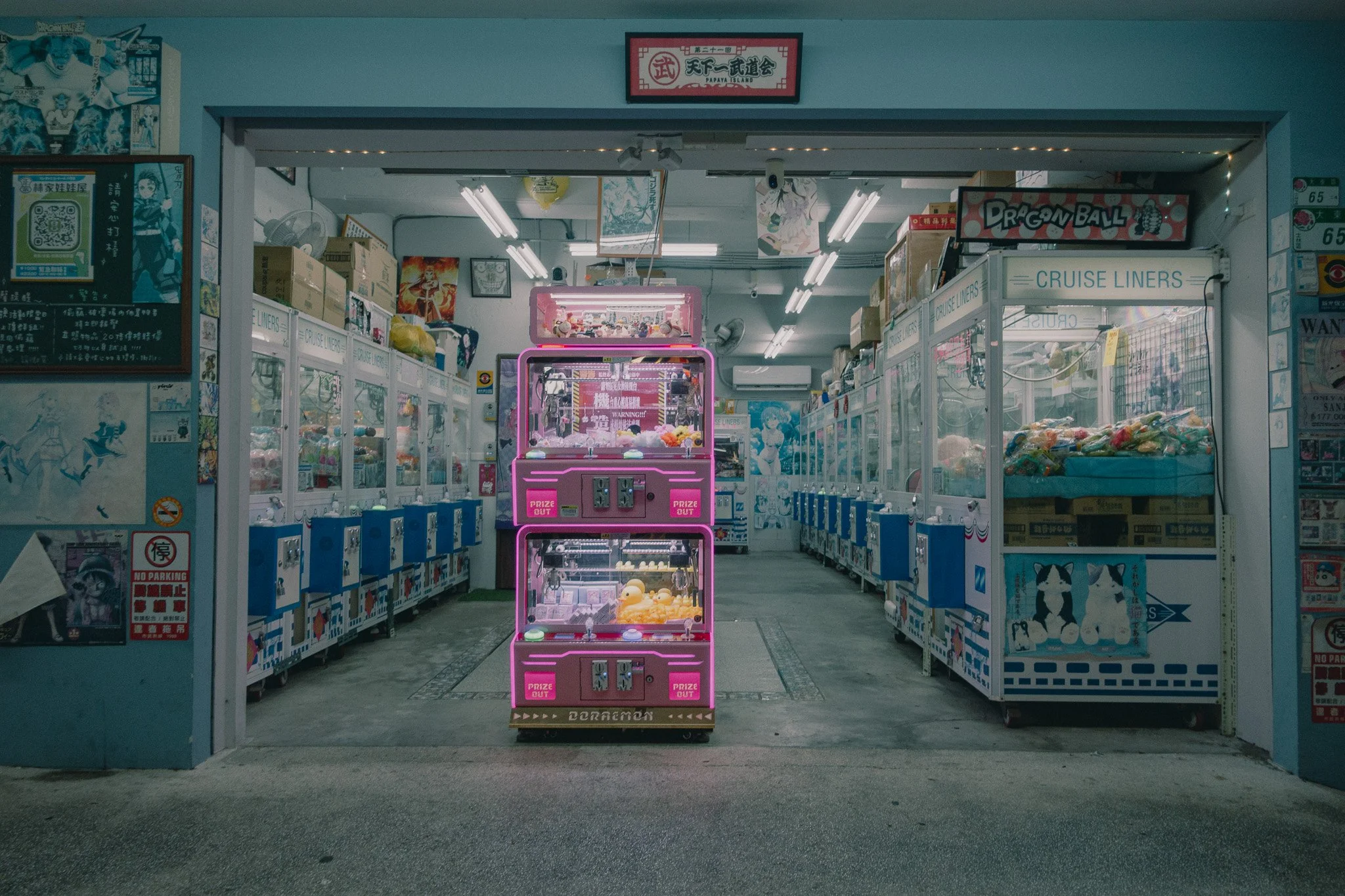 An arcade filled with claw machines, featuring colorful plush toys and neon pink signage, in a space decorated with posters and gaming memorabilia.