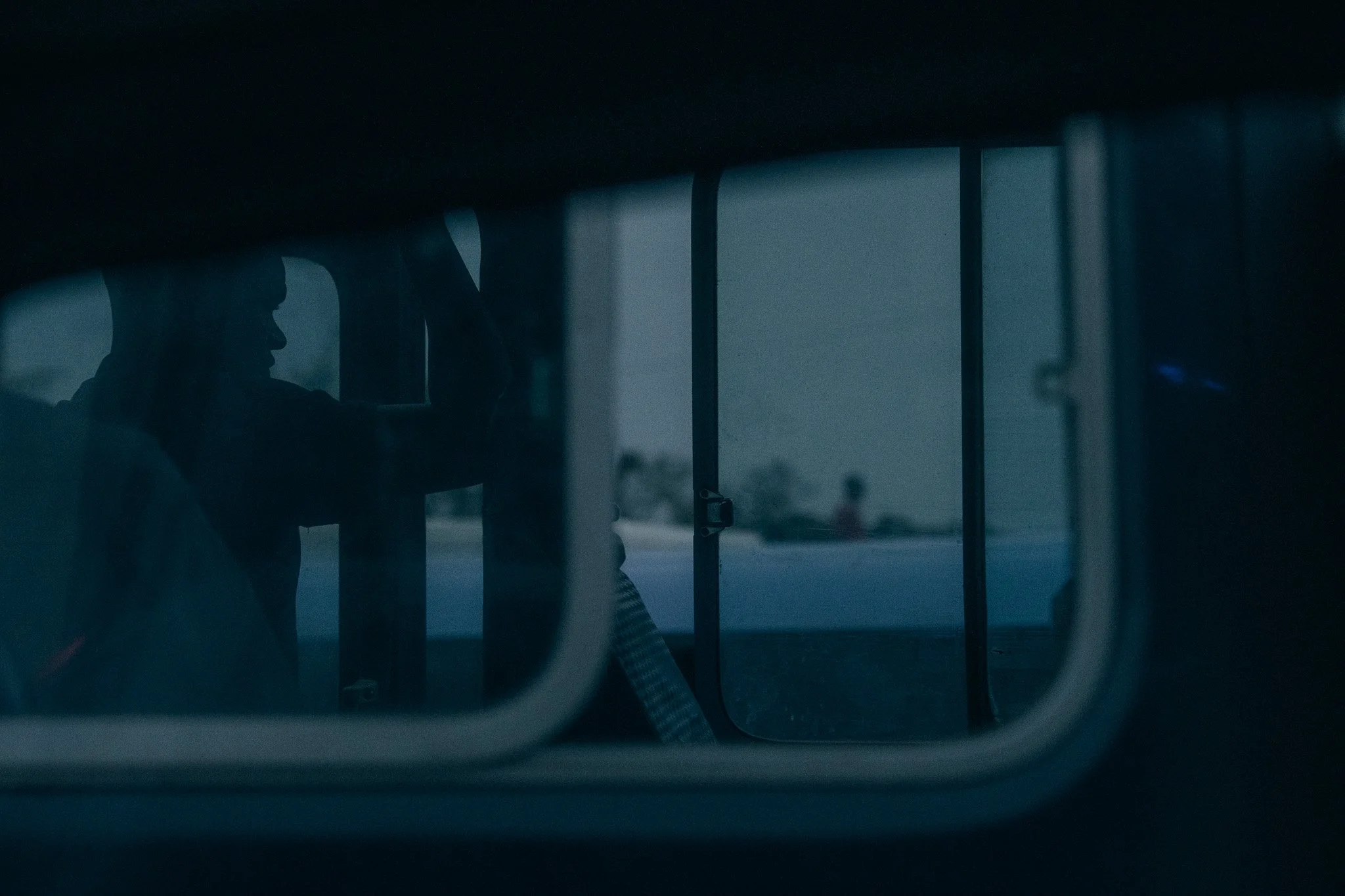 A person with glasses and short hair sitting inside a vehicle, seen through the window with bars, during twilight or late evening.