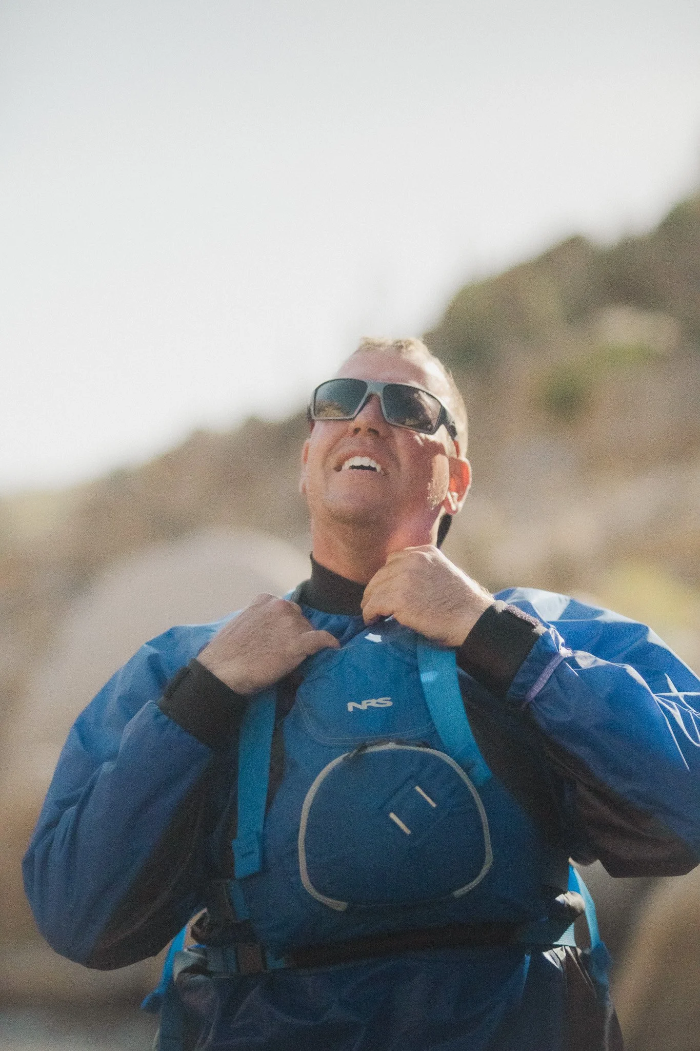 A man in a blue jacket and sunglasses adjusting his collar outdoors during daytime.