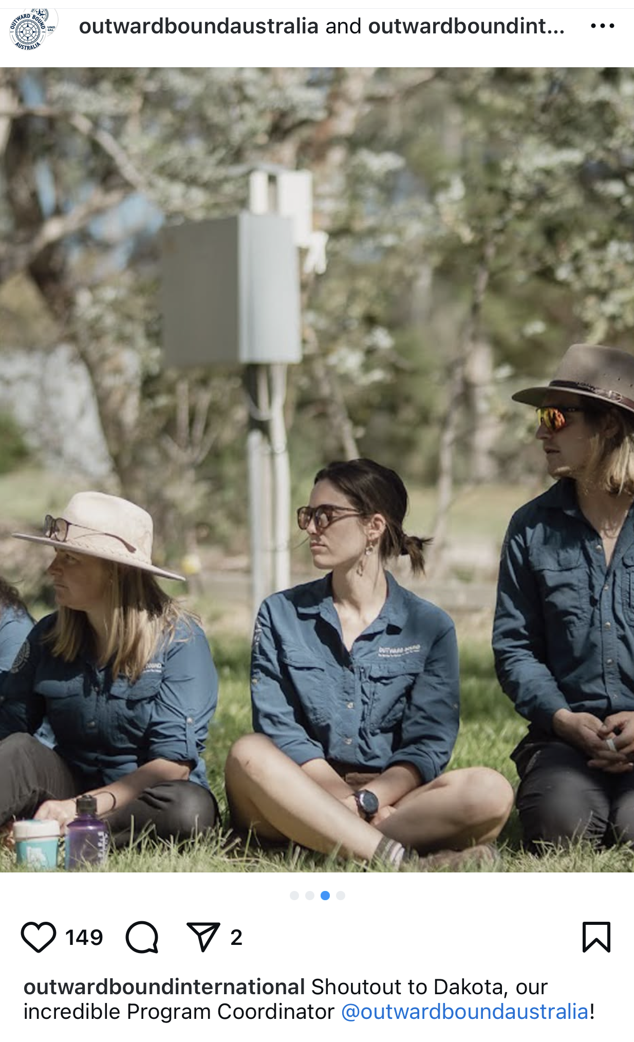 Group of women sitting outdoors on grass, wearing outdoor clothing and hats, with a background of trees and communication equipment.