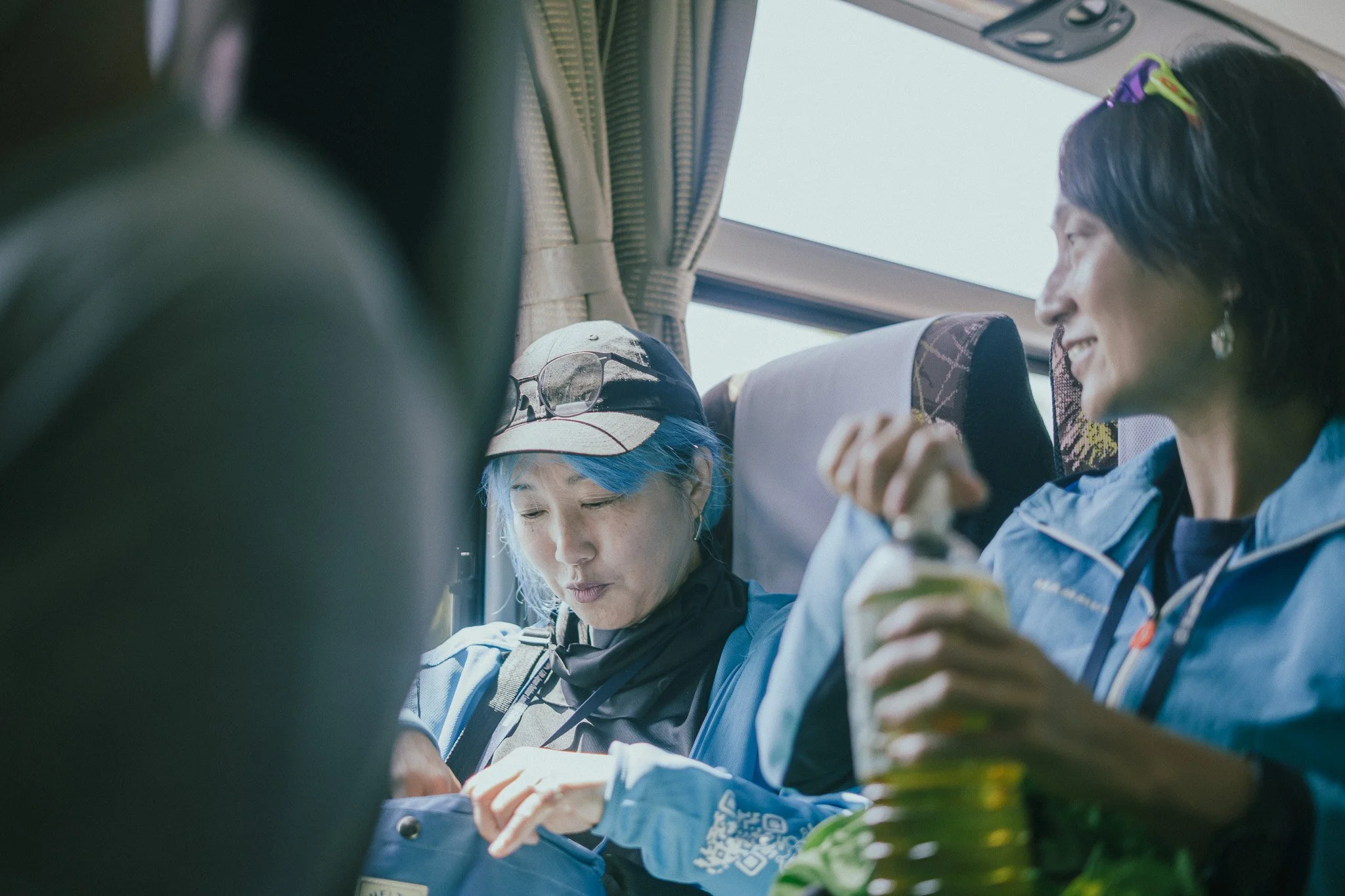 Three women, two with dark hair and one with light blue hair, sitting closely together on a bus, eating snacks and engaging in a conversation. They are wearing outdoor jackets and sunglasses are placed on the light blue-haired woman's hat.