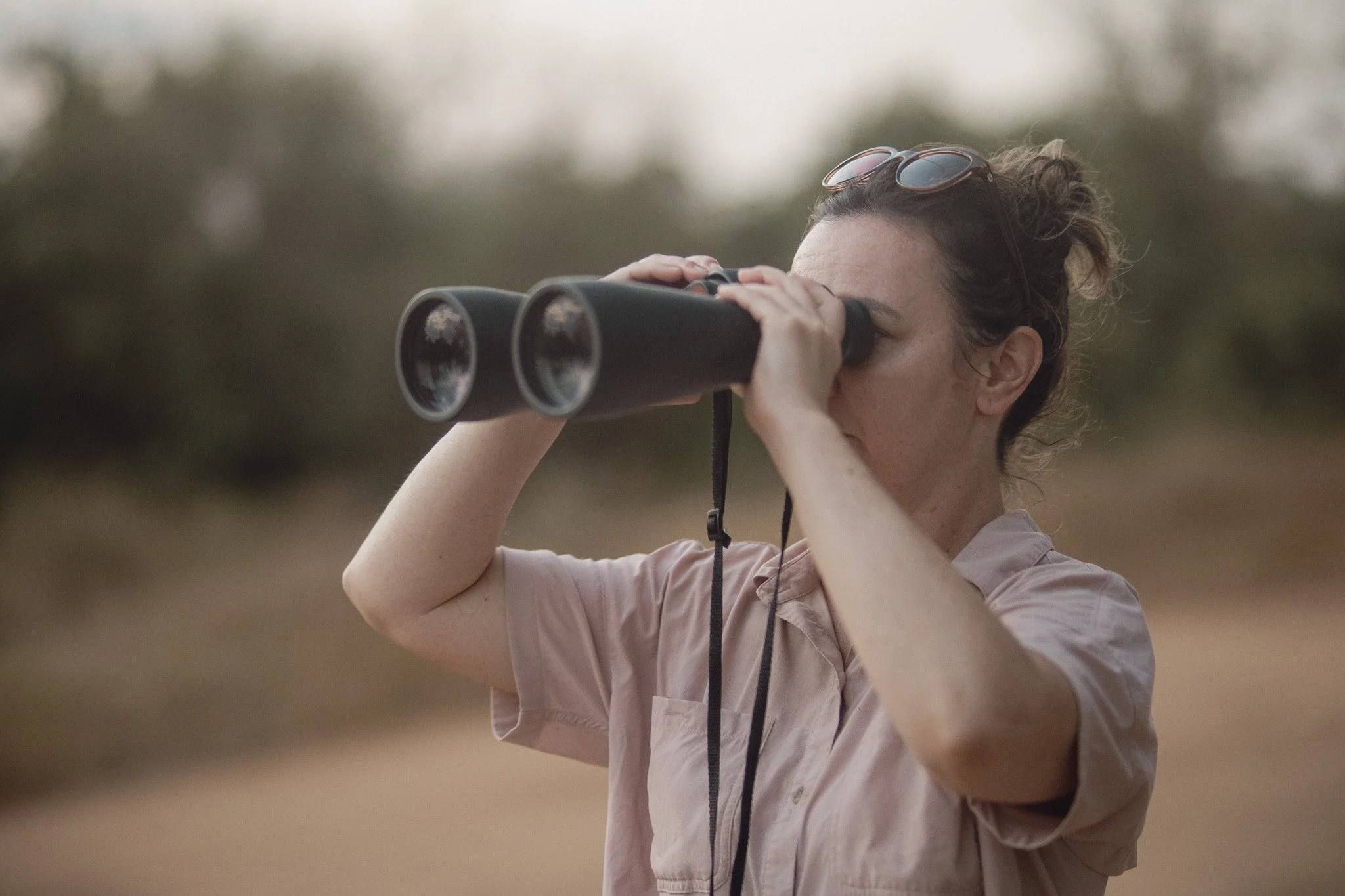 A person with curly hair is looking through binoculars outdoors, possibly on a nature hike or birdwatching, with a blurred background of trees.