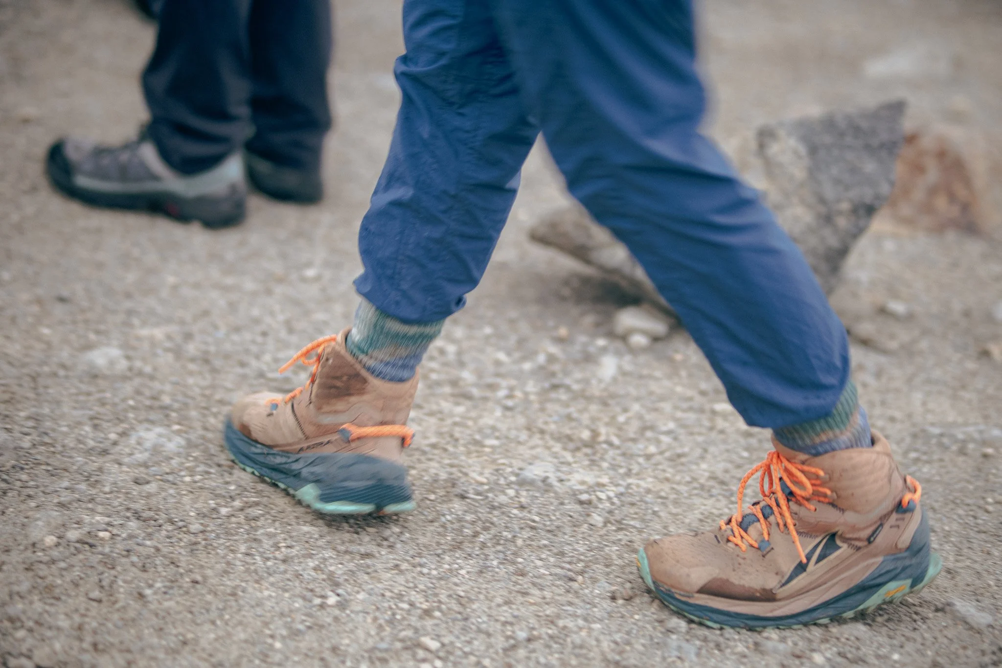 Child wearing tan hiking shoes with orange laces, navy pants, and colorful socks, walking on a gravel trail.