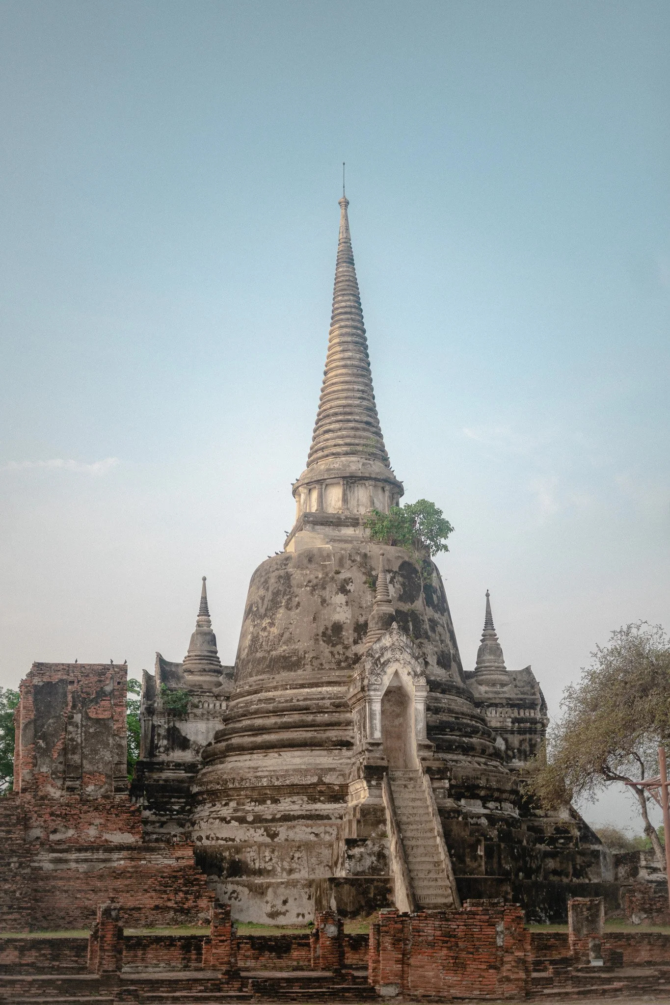 An ancient stone temple with a tall, pointed spire, surrounded by smaller structures and brick walls, with trees and a cloudy sky in the background.