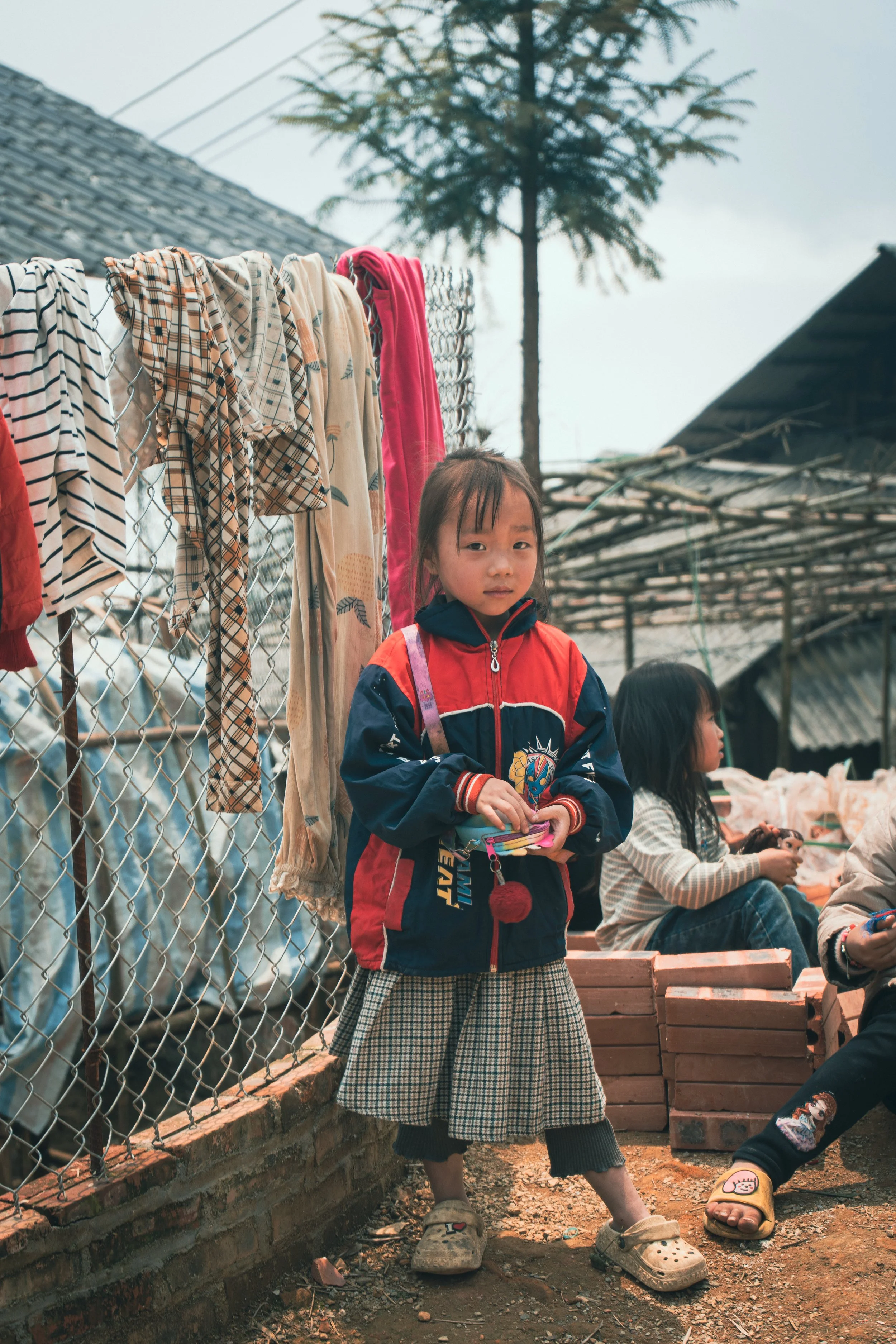 A young girl in a red and navy jacket, plaid skirt, and Crocs shoes standing outdoors with a fence and laundry hanging behind her. Other children are seated nearby against a background of trees and buildings.