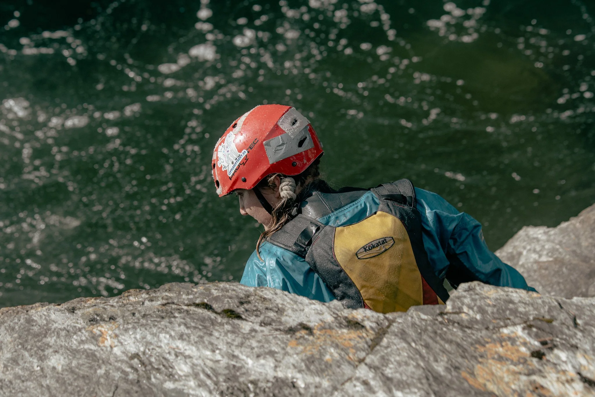 Person wearing a red helmet and a blue jacket climbing a rocky surface near a body of water.