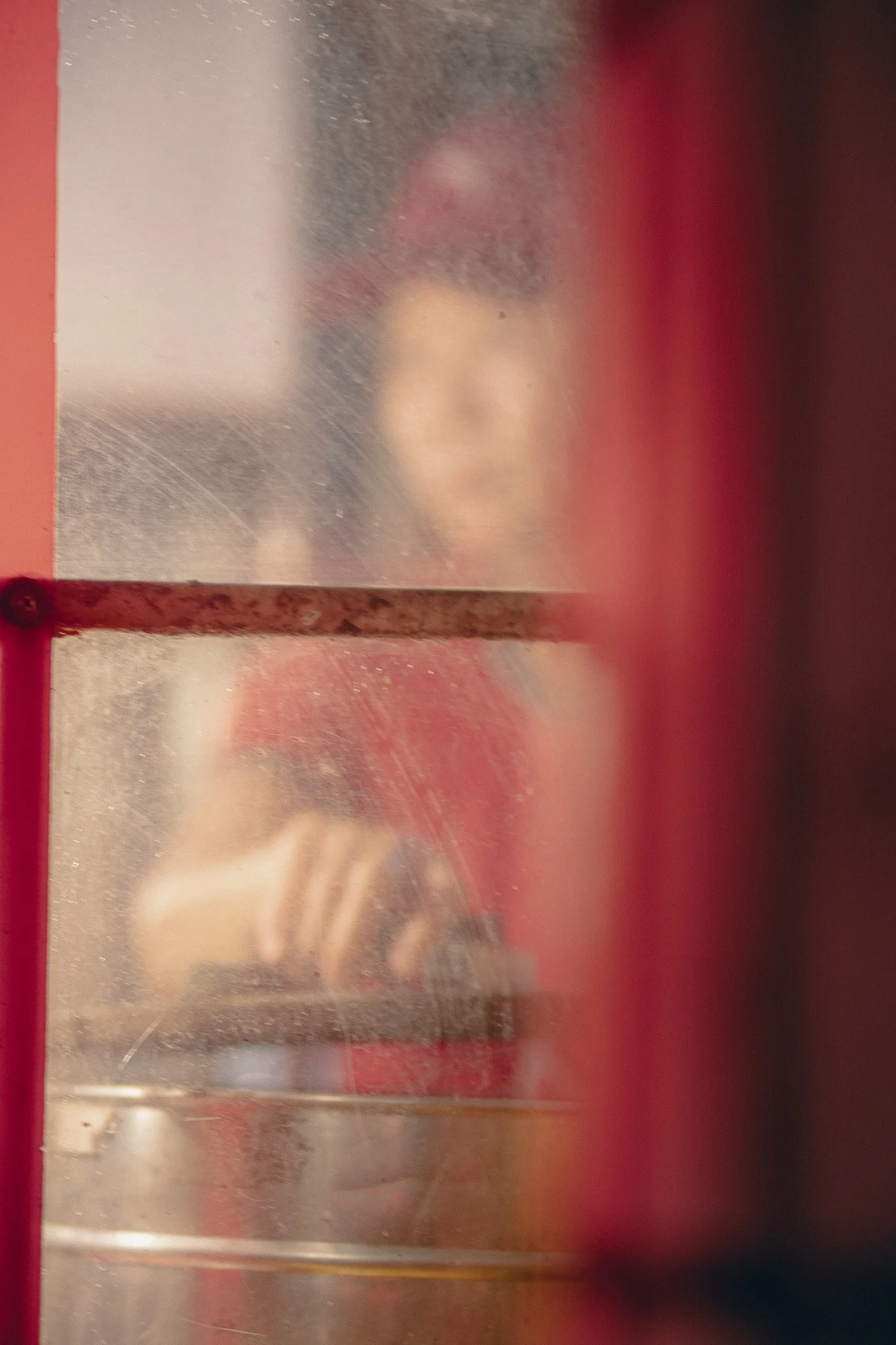 Person taking a photo through a frosted glass window with red frame, showing a blurred reflection of their face and hand.