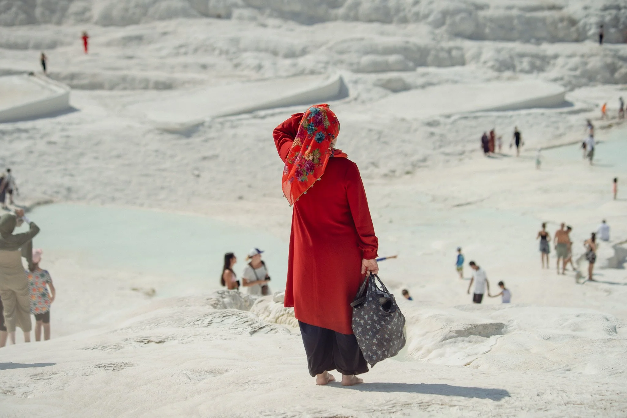 Woman in a red coat and colorful headscarf walking barefoot on a snow-covered landscape, carrying a black bag, with other people in the background.