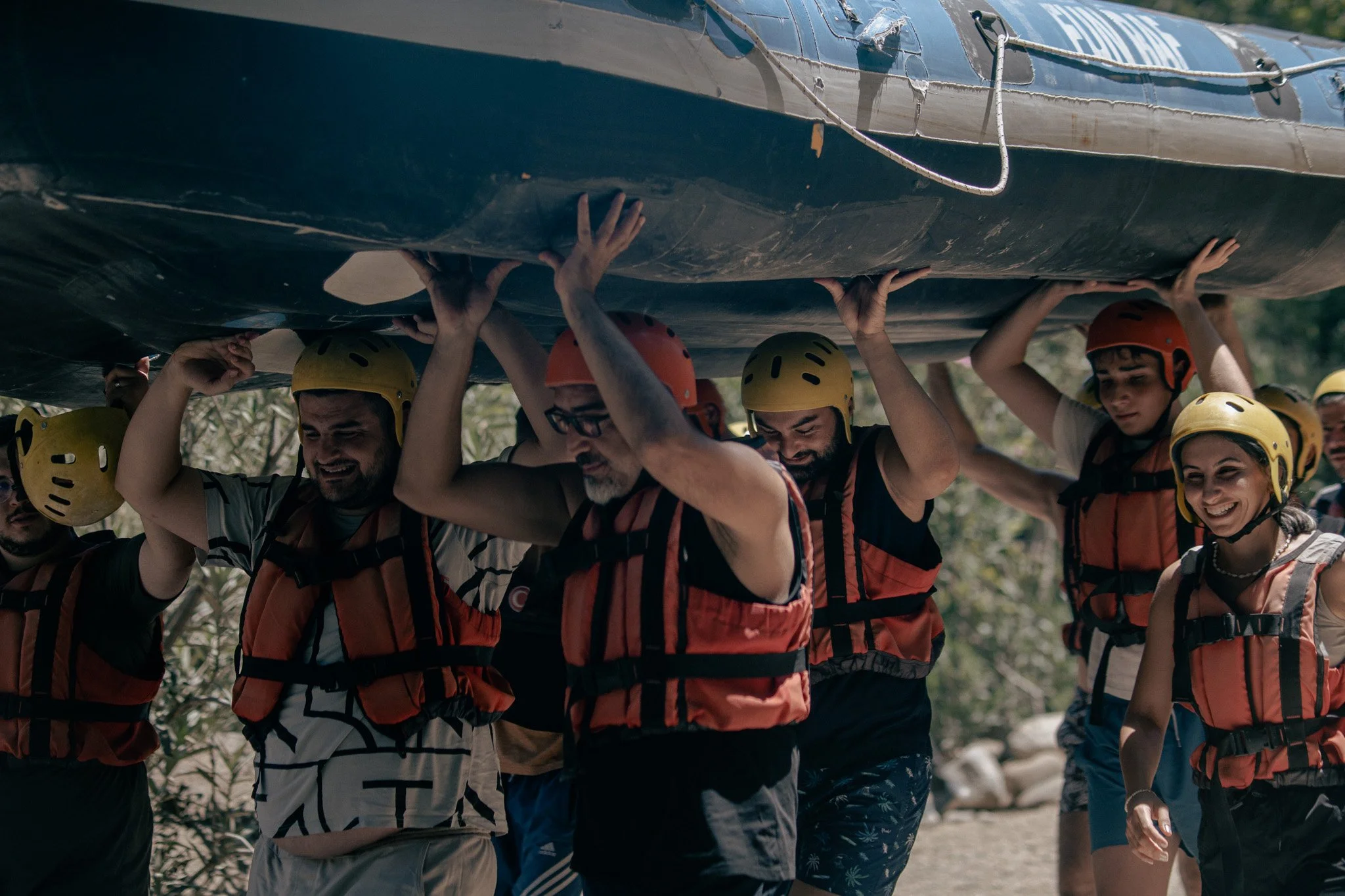 A group of people wearing life jackets and helmets, carrying a boat on their shoulders outdoors.