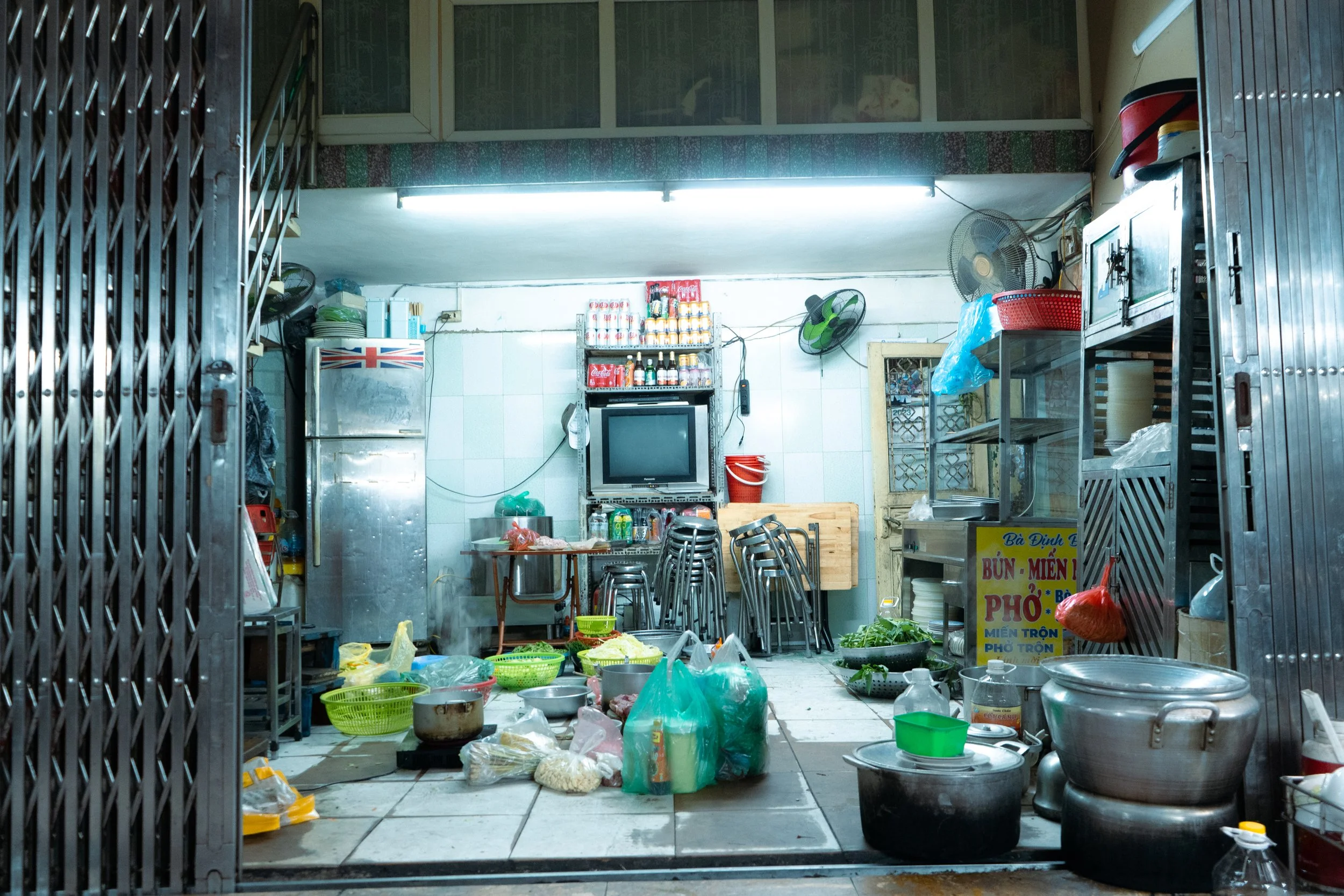 Interior of a small, cluttered eatery with stacked chairs, a television, various containers, and cooking supplies scattered on the floor and shelves.
