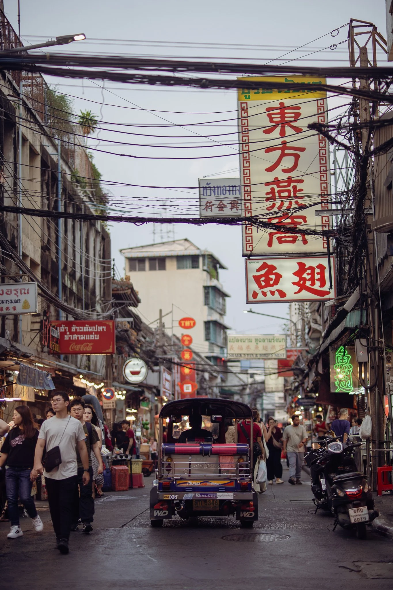 Busy street market scene with people walking, chao tuktuk vehicle in the center, and multiple signs in Asian scripts hanging above, with electrical wires overhead.