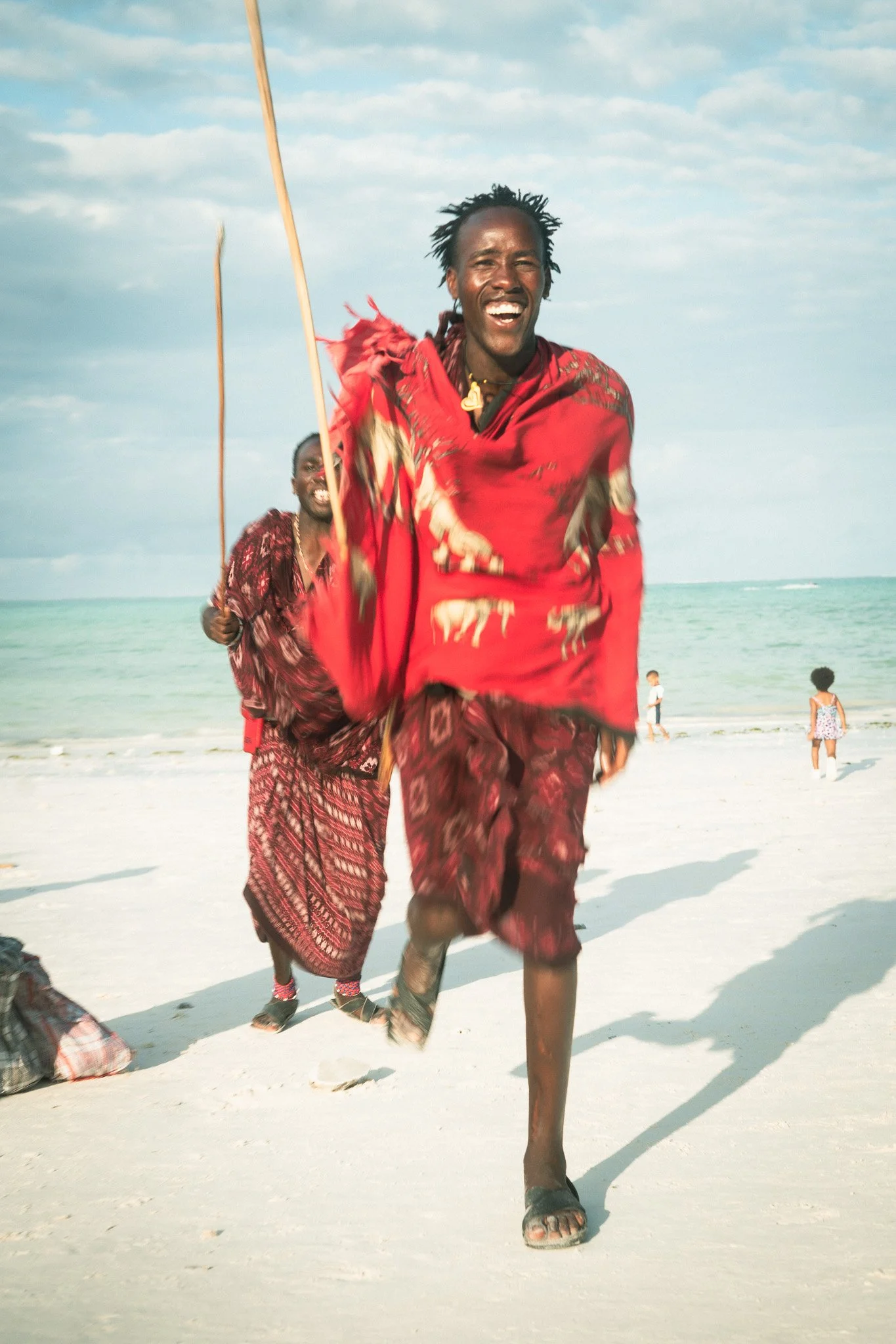 Two men in traditional clothes running happily on the beach near the ocean, with children playing in the background.