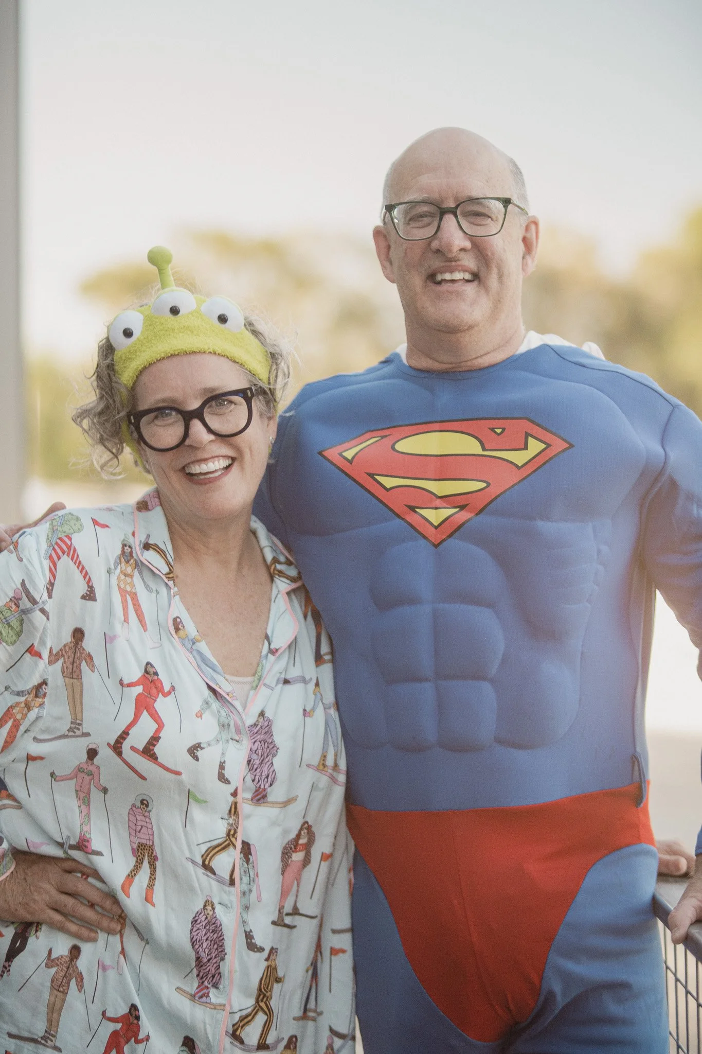 A cheerful woman wearing a green alien headband and glasses stands next to a man in a Superman costume. They are outdoors, smiling for the camera.