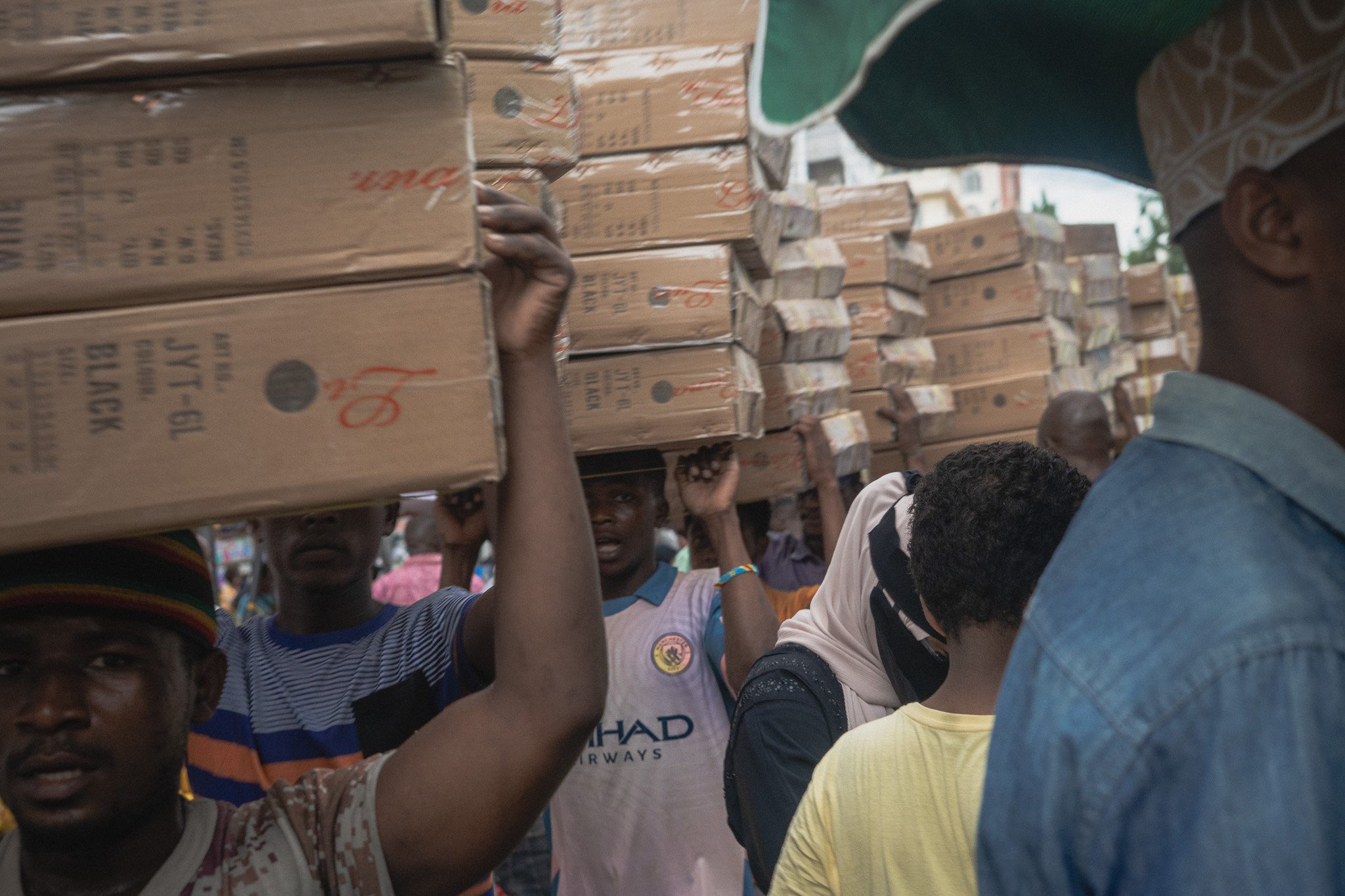 People carrying stacked boxes on their heads in a crowded marketplace or street.