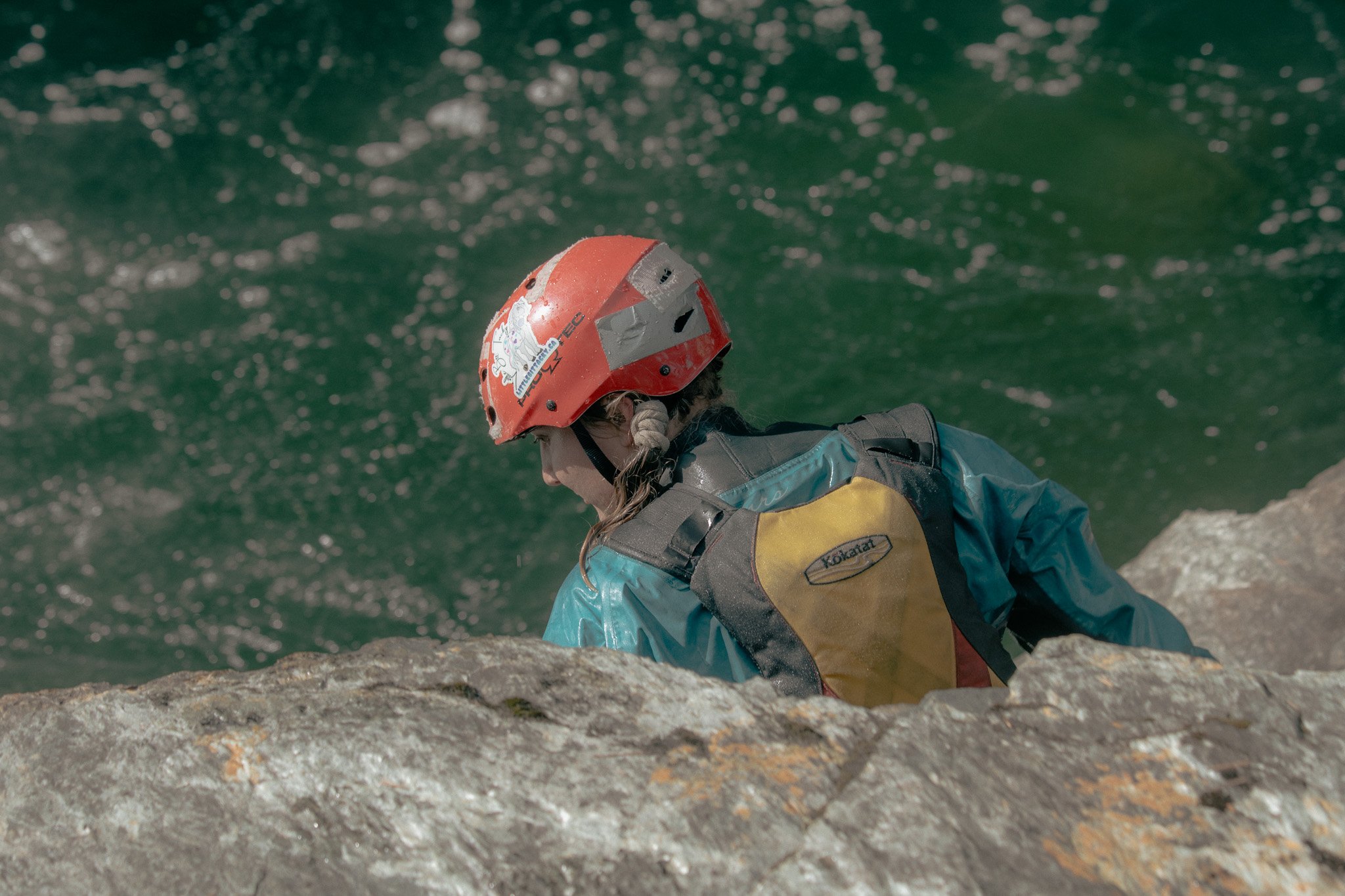 A person wearing a red helmet, waterproof jacket, and vest sitting on a rocky surface near a greenish river, with water splashing around.