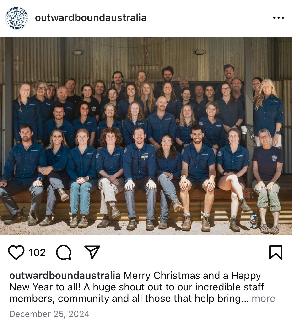 Group photo of staff members wearing matching blue shirts, posed together in front of a rustic wooden and metal background.