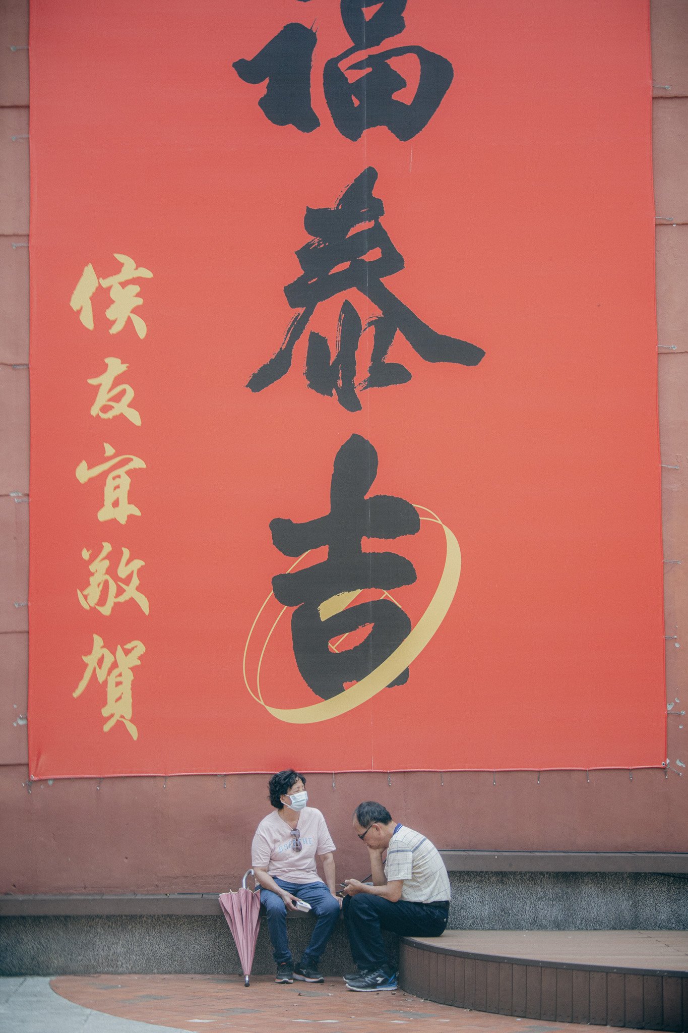 Two people sitting on a bench beneath a large red banner with black and gold Chinese characters. One person is wearing a face mask and holding a pink umbrella, and the other is looking at a mobile phone.