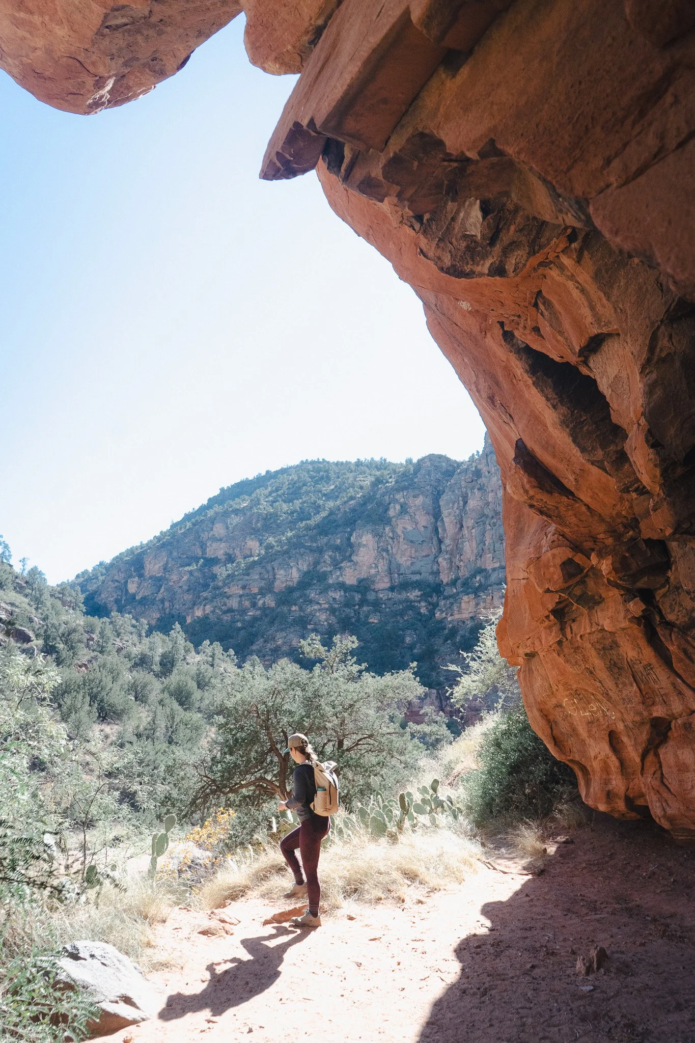 A woman with a backpack hiking in a canyon with large red rock formations and desert vegetation.