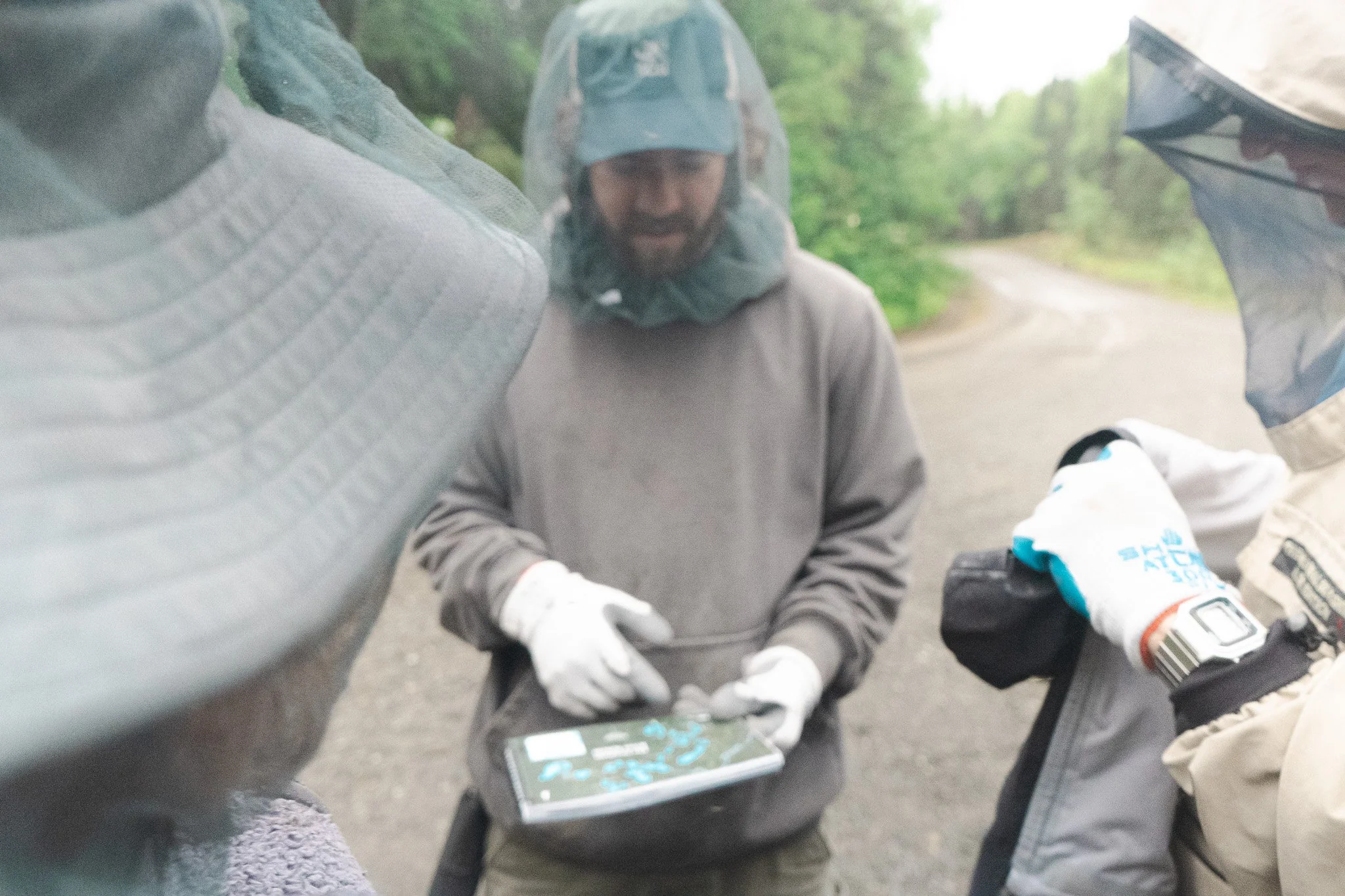 Three people outdoors viewing a map on a handheld device, wearing protective gear including gloves, hats, and hoods, with a green wooded background.
