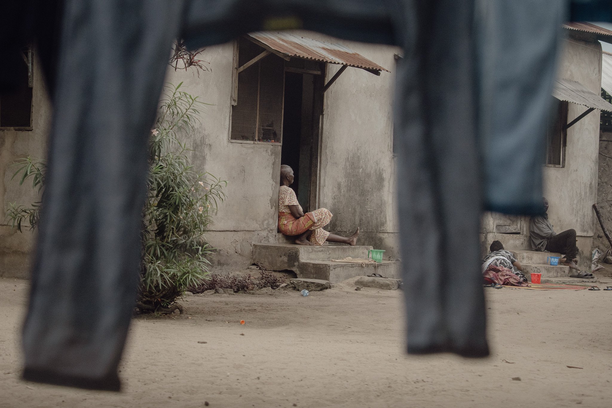 An elderly person sitting on the steps of a worn house, seen through hanging clothing in a rural setting.