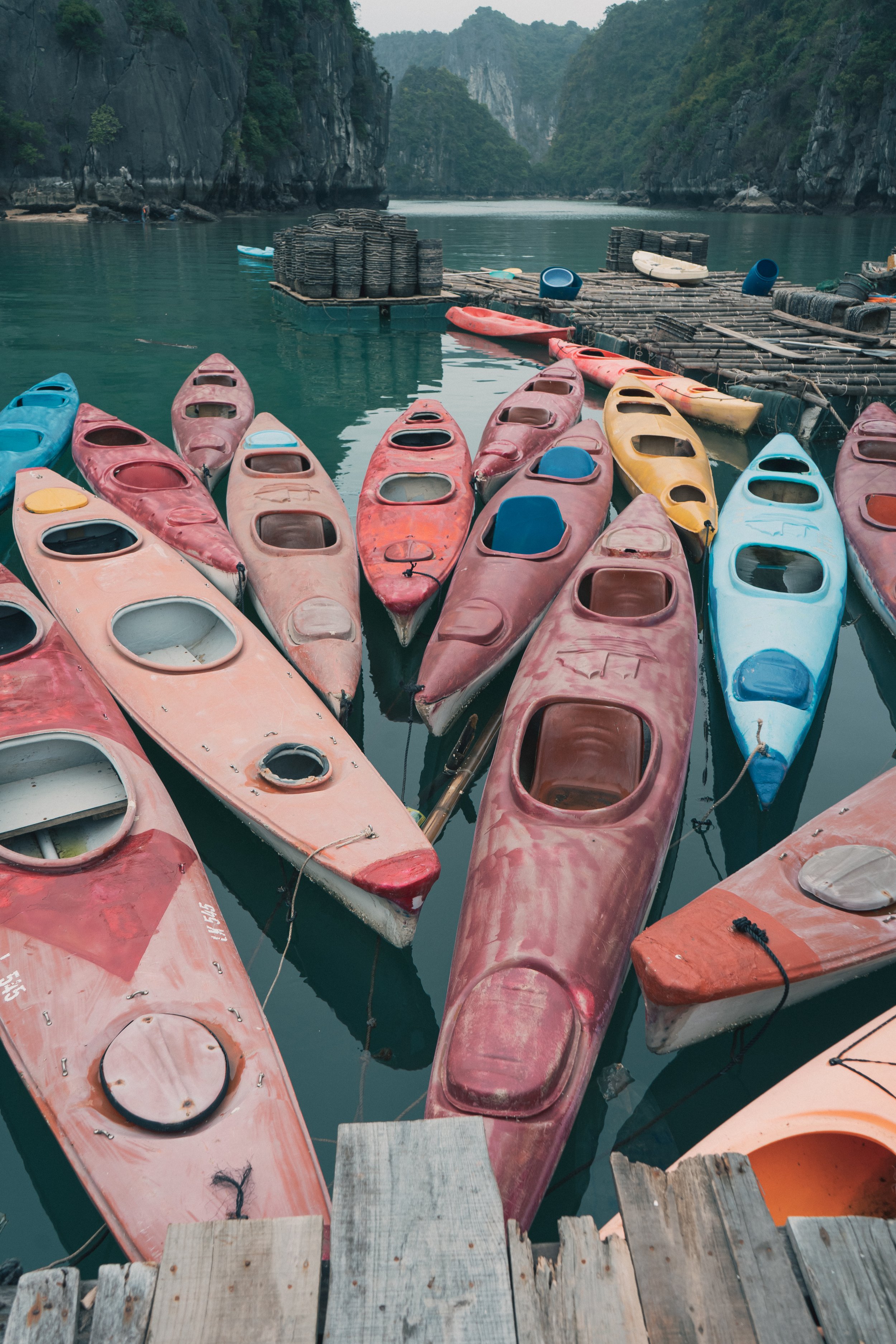 Multiple colorful kayaks docked at a wooden pier on a calm river, with forested hills in the background.