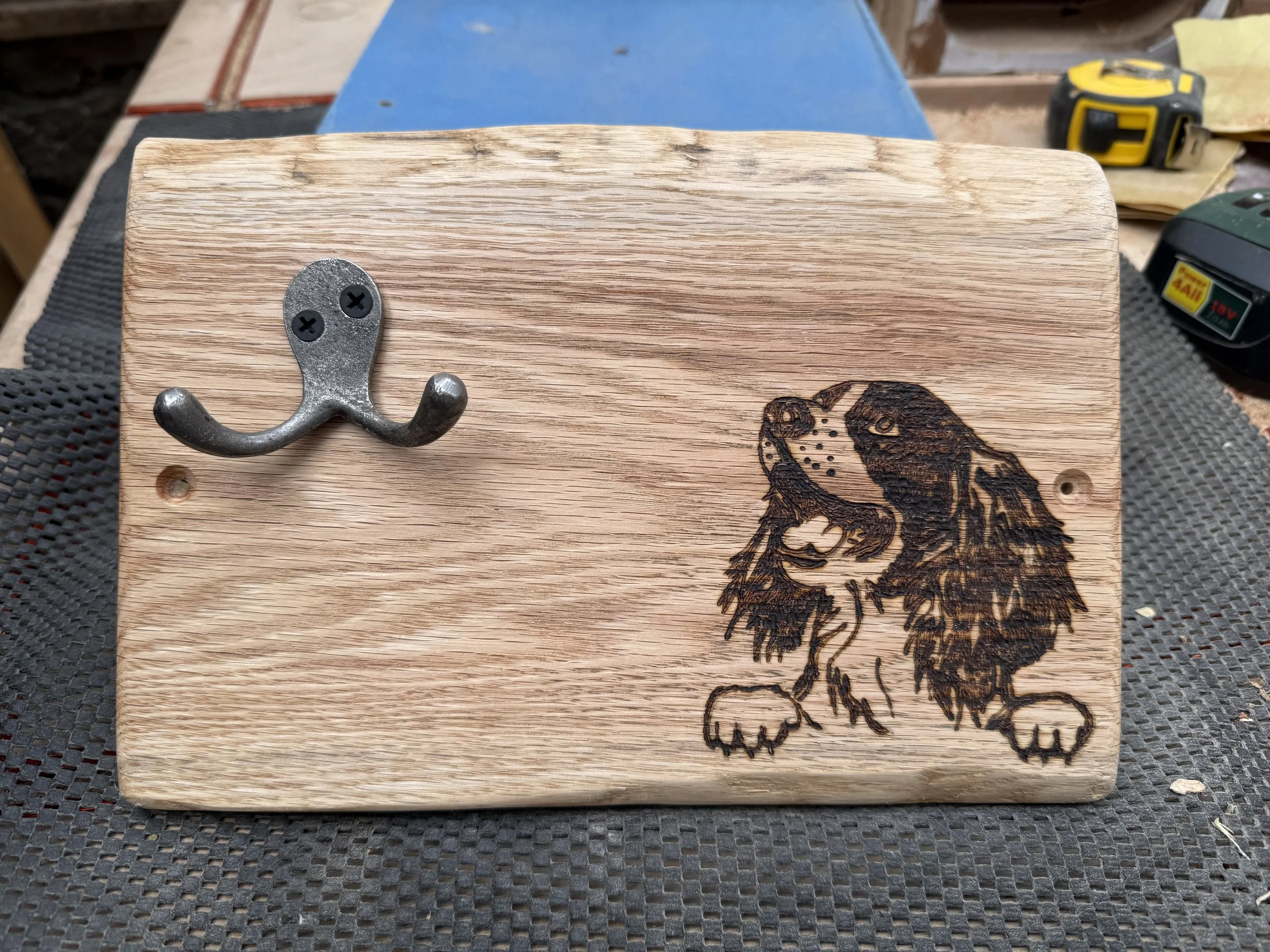 A wooden block with a metal hook attached, and a partially burned image of a dog burned into the wood, along with a tape measure and other tools in the background.