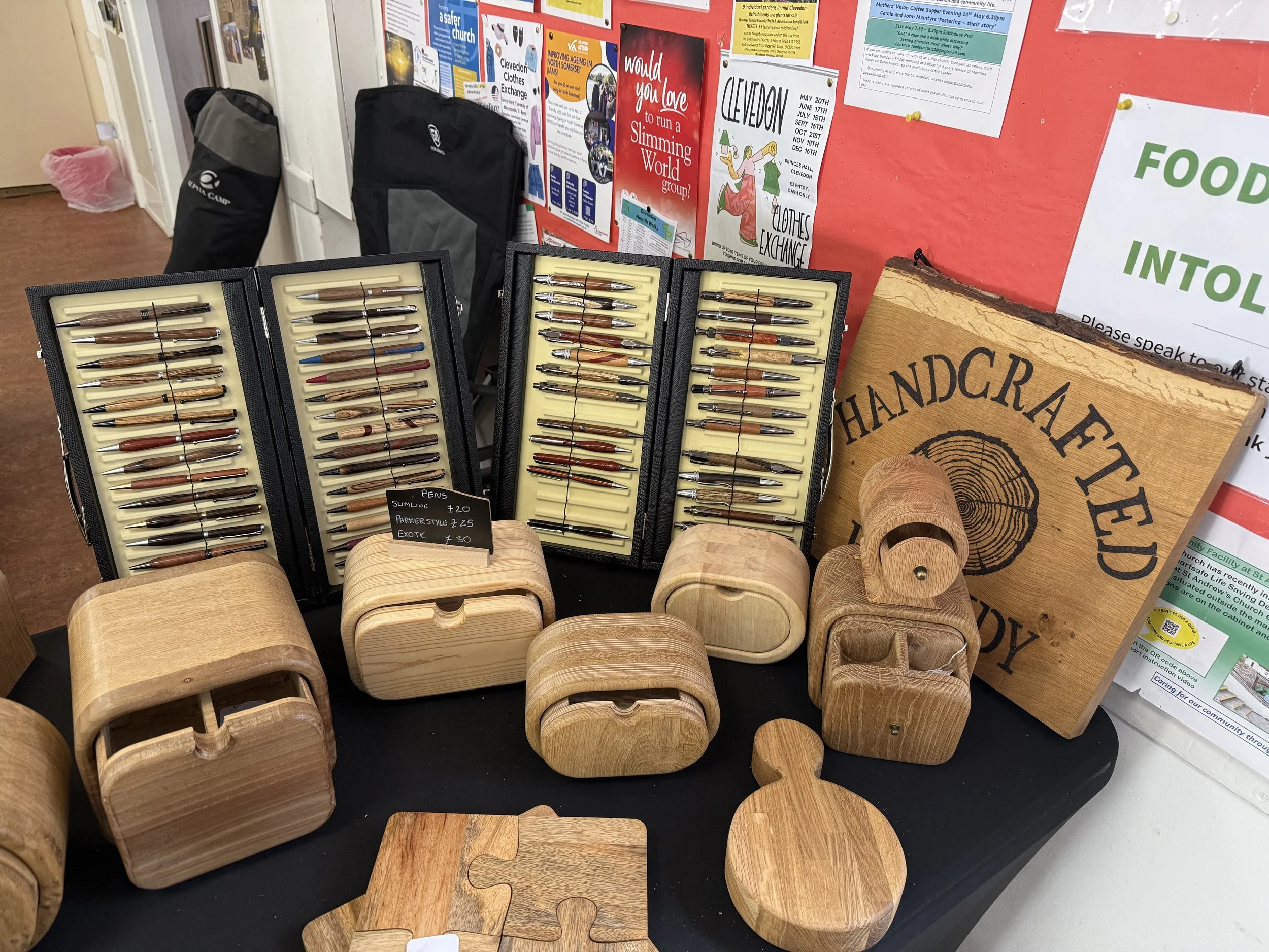 Display of wooden pens in cases and wooden animal-shaped stools at a craft fair, with posters and notices on a red background wall behind them.