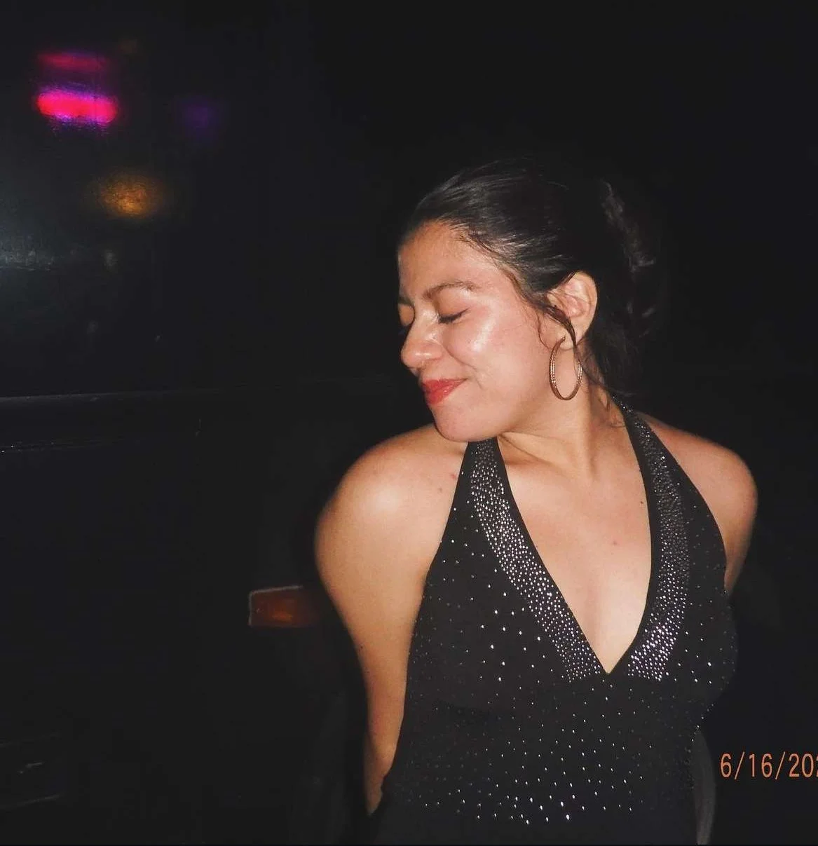 A young woman with dark hair in a bun, wearing hoop earrings and a black, sparkly halter dress, smiles with her eyes closed against a dark background.