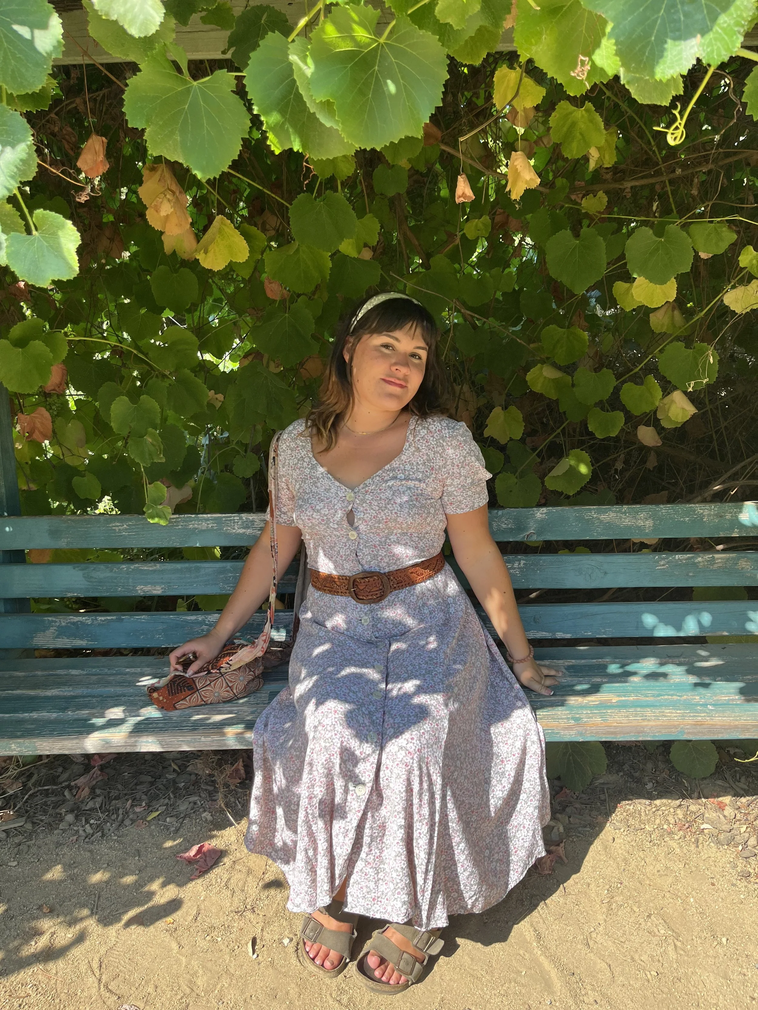 Woman in floral dress sitting on a green wooden bench, surrounded by green vine leaves and shade