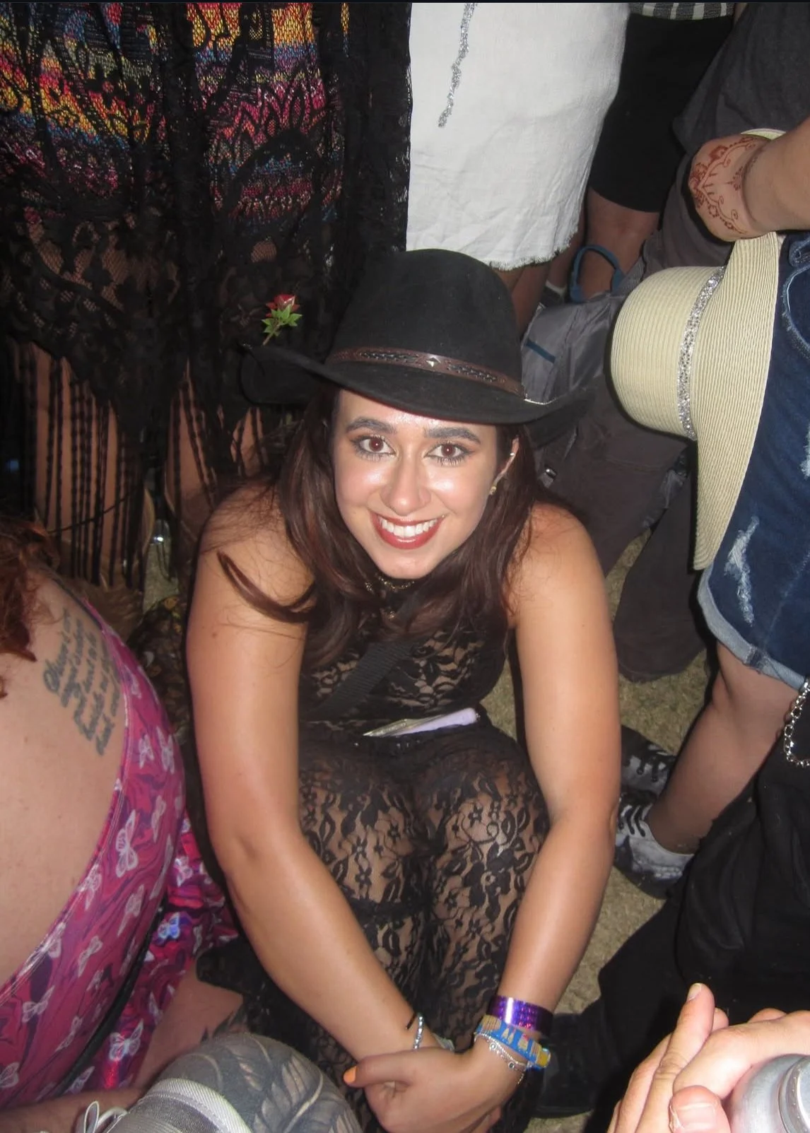 A woman smiling and sitting on the ground, wearing a black hat, a black lace dress, and colorful wristbands, surrounded by people at a crowded event.