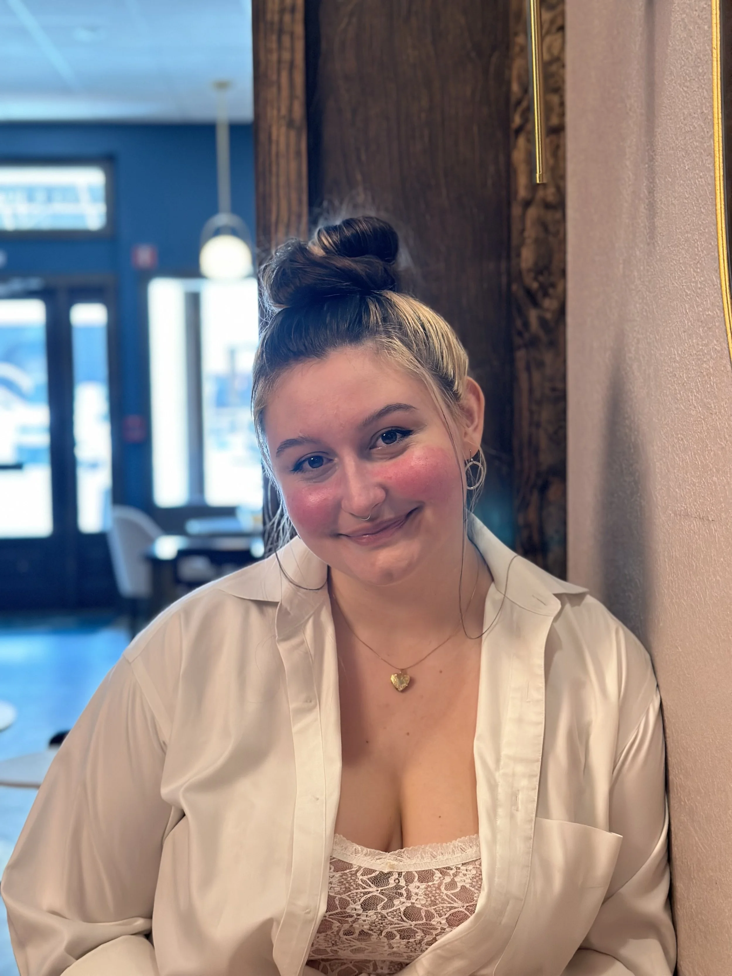 Young woman with a top bun hairstyle, wearing a beige satin jacket over a pink lace top, sitting inside a restaurant or cafe. She has a gold heart necklace, hoop earrings, and is smiling softly.