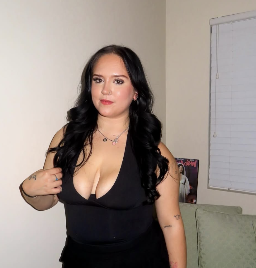 A woman with long black hair wearing a sparkly silver top, black shorts, and knee-high black boots standing on a balcony at night, with her left hand on the railing and her right hand touching her neck.