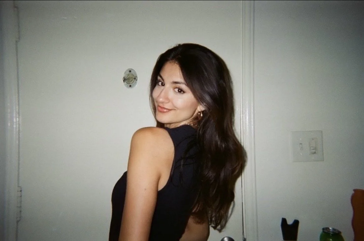 A woman with long dark hair and a black sleeveless top looking over her shoulder and smiling at the camera in a room with white walls.
