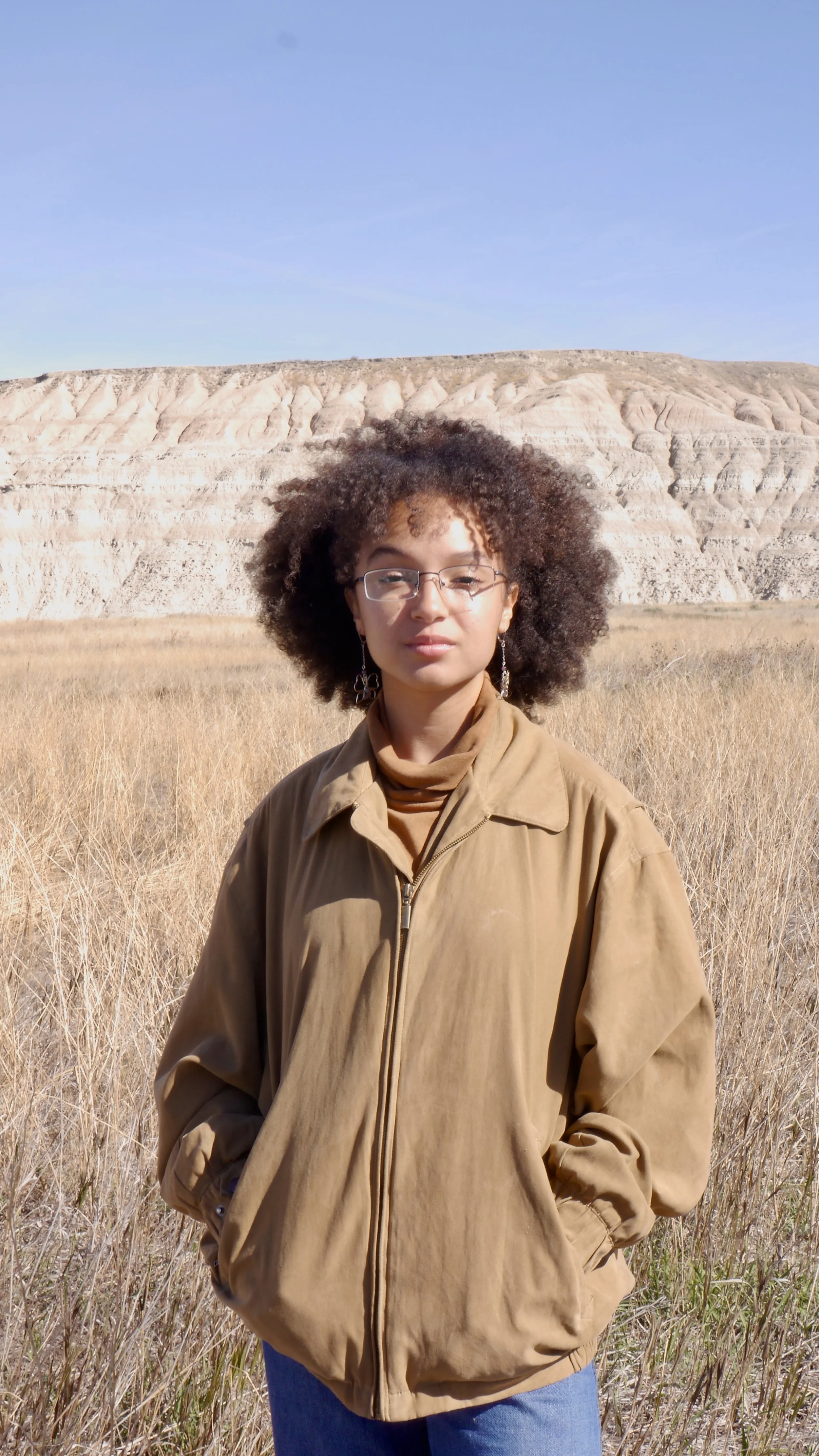 A woman with curly hair and glasses standing in a dry grassy field with a layered rock formation in the background under a clear blue sky.