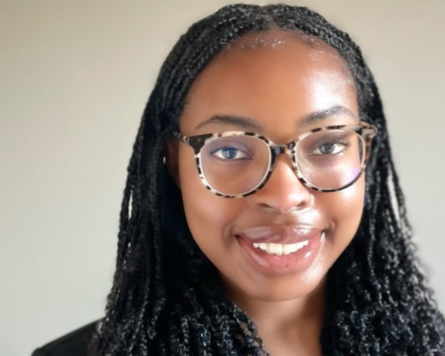 Close-up of a smiling woman with black twist braids, wearing tortoiseshell glasses and a black blazer against a beige background.