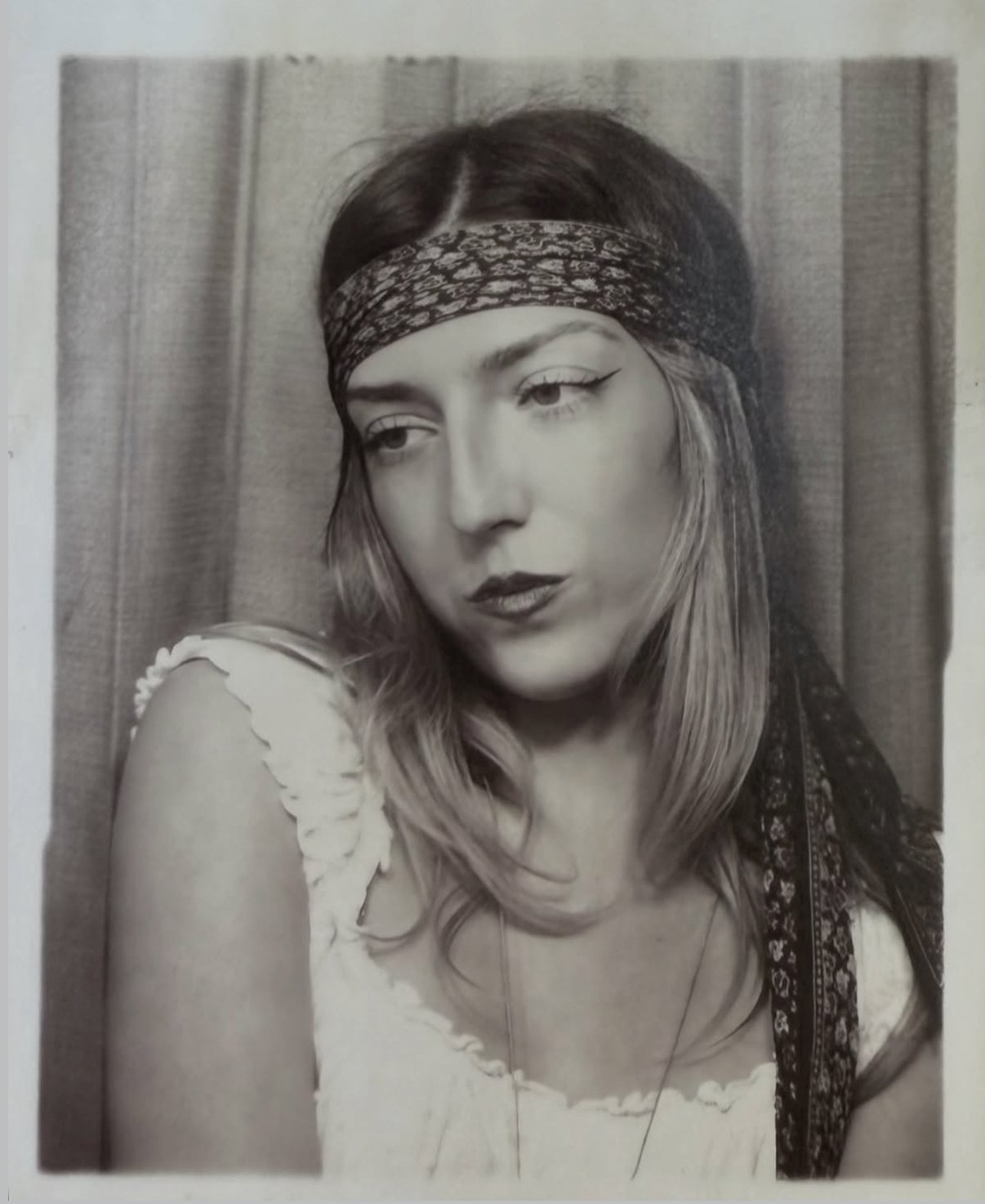 A woman with short blonde hair, makeup, and a septum piercing, sitting on a ledge near a pool with blue mosaic tiles, wearing a black sleeveless top and camouflage shorts.