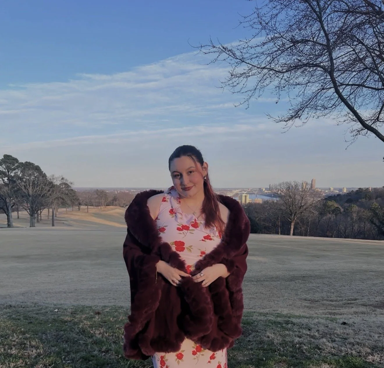 Young woman with a top bun hairstyle, wearing a beige satin jacket over a pink lace top, sitting inside a restaurant or cafe. She has a gold heart necklace, hoop earrings, and is smiling softly.