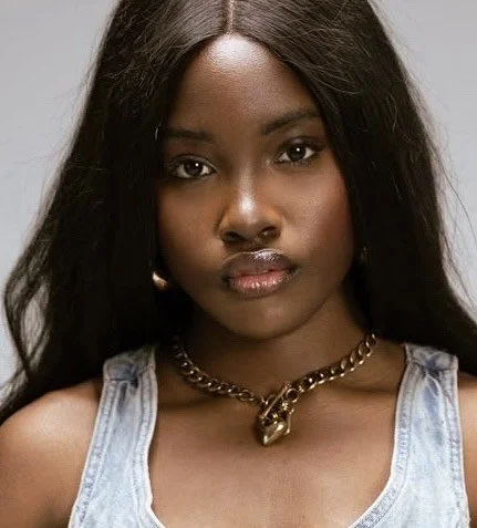 Portrait of a young Black woman with long black hair, wearing a denim top and a chunky gold chain necklace with a skull pendant.